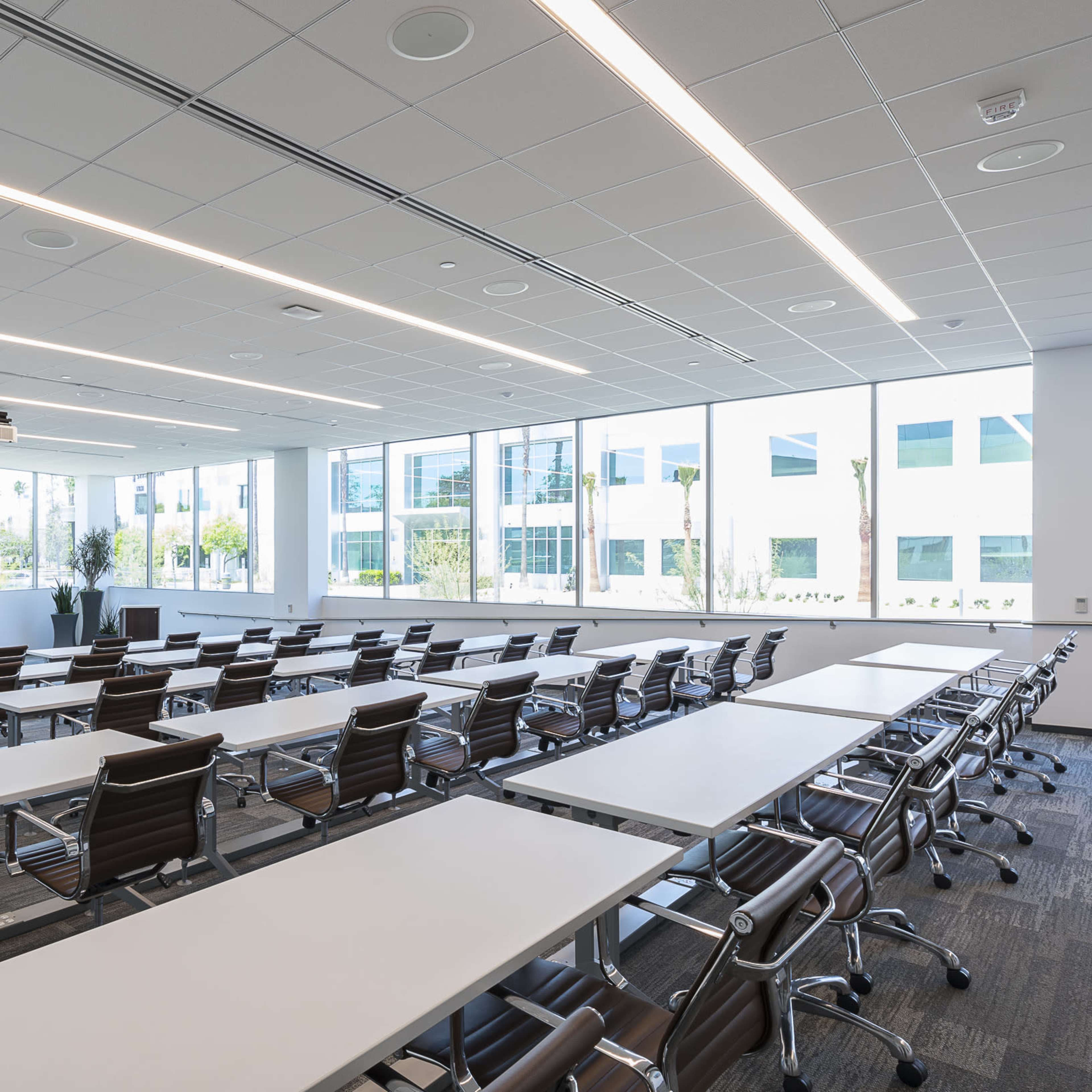 A modern conference room features rows of tables and chairs arranged toward a projection screen at the front, with large windows providing natural light.