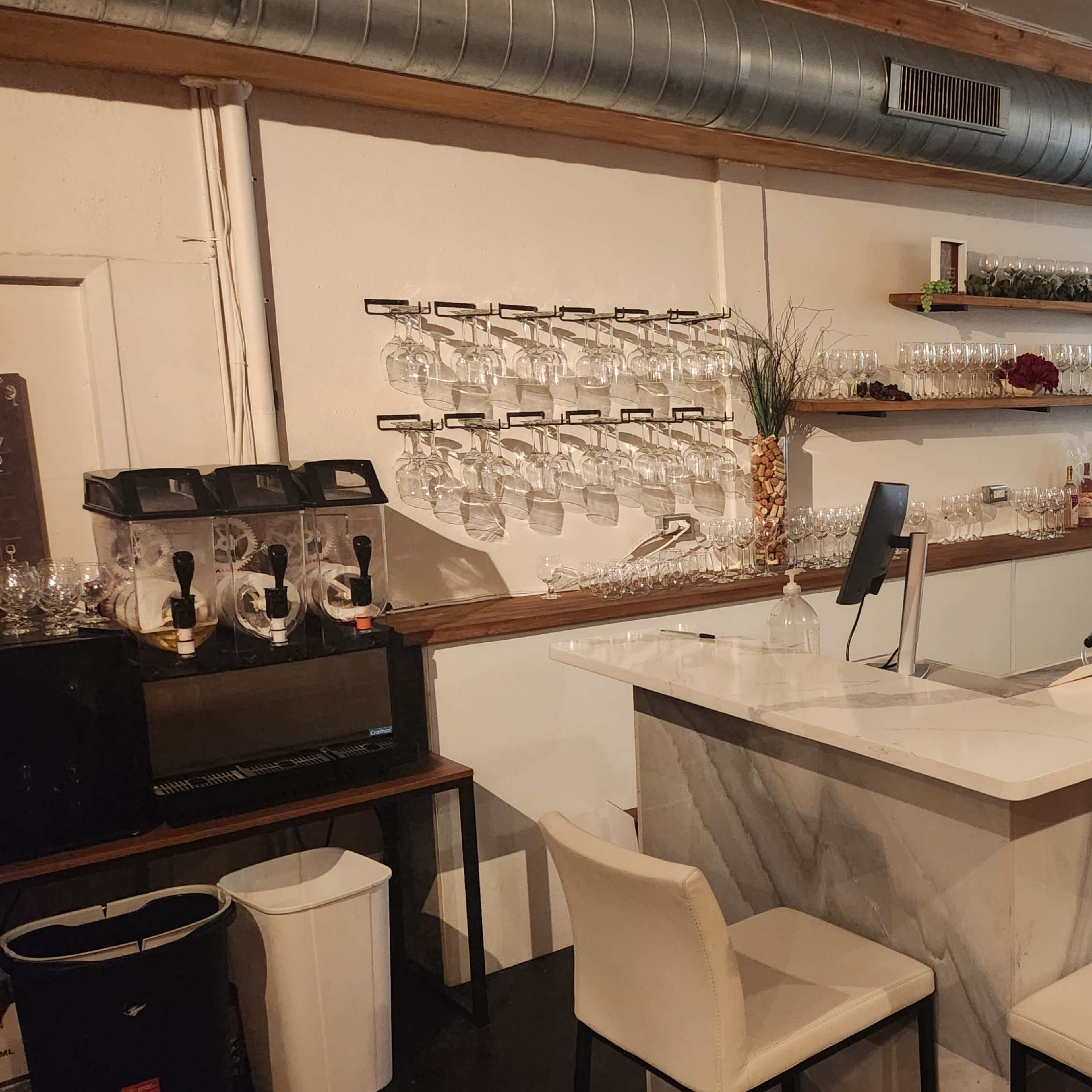 A modern bar area with a marble countertop, tall chairs, and a wall of hanging wine glasses, illuminated by overhead lighting.