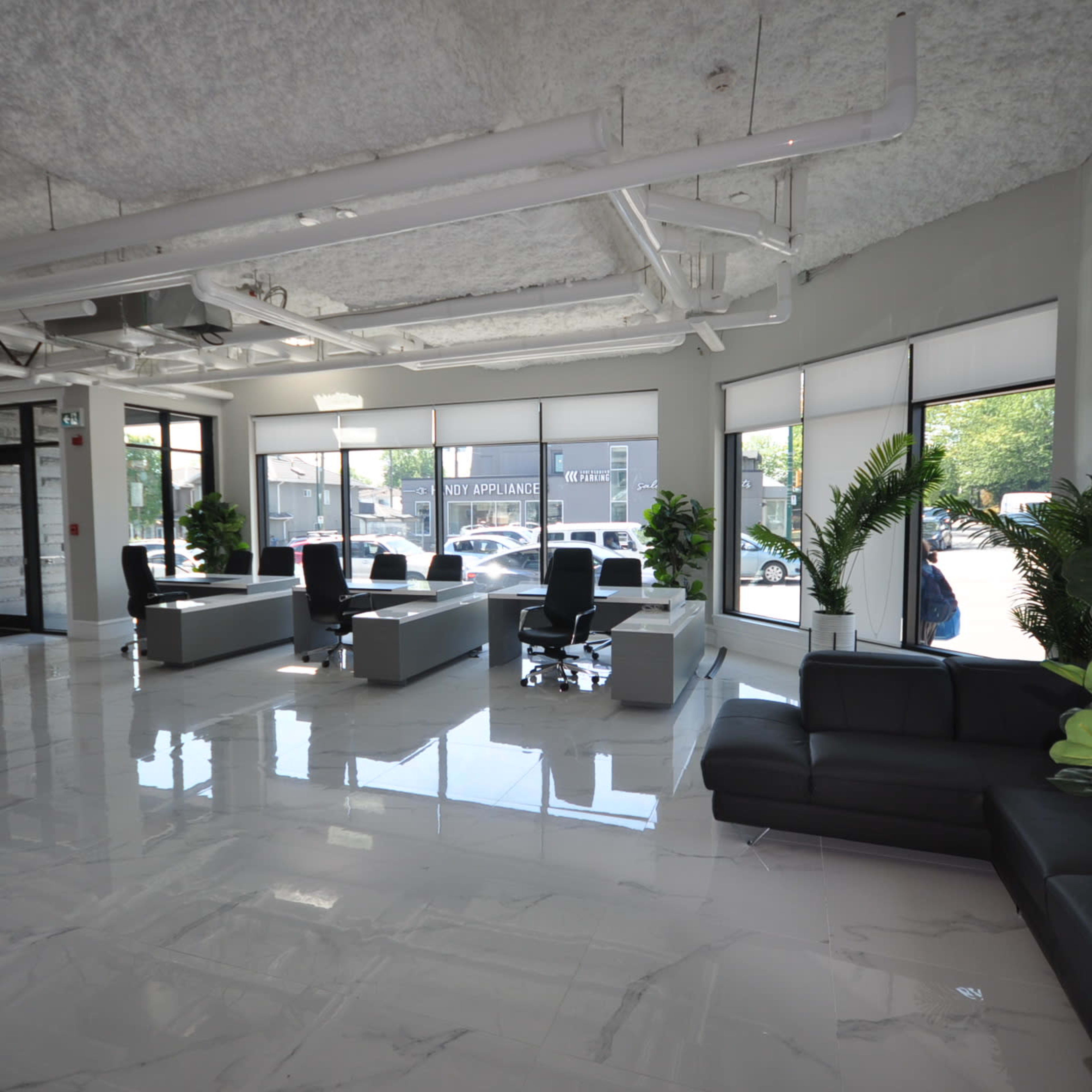 A modern lobby area with gray walls, polished white tile flooring, and multiple seating arrangements, including sofas and office chairs, surrounded by large windows and potted plants.