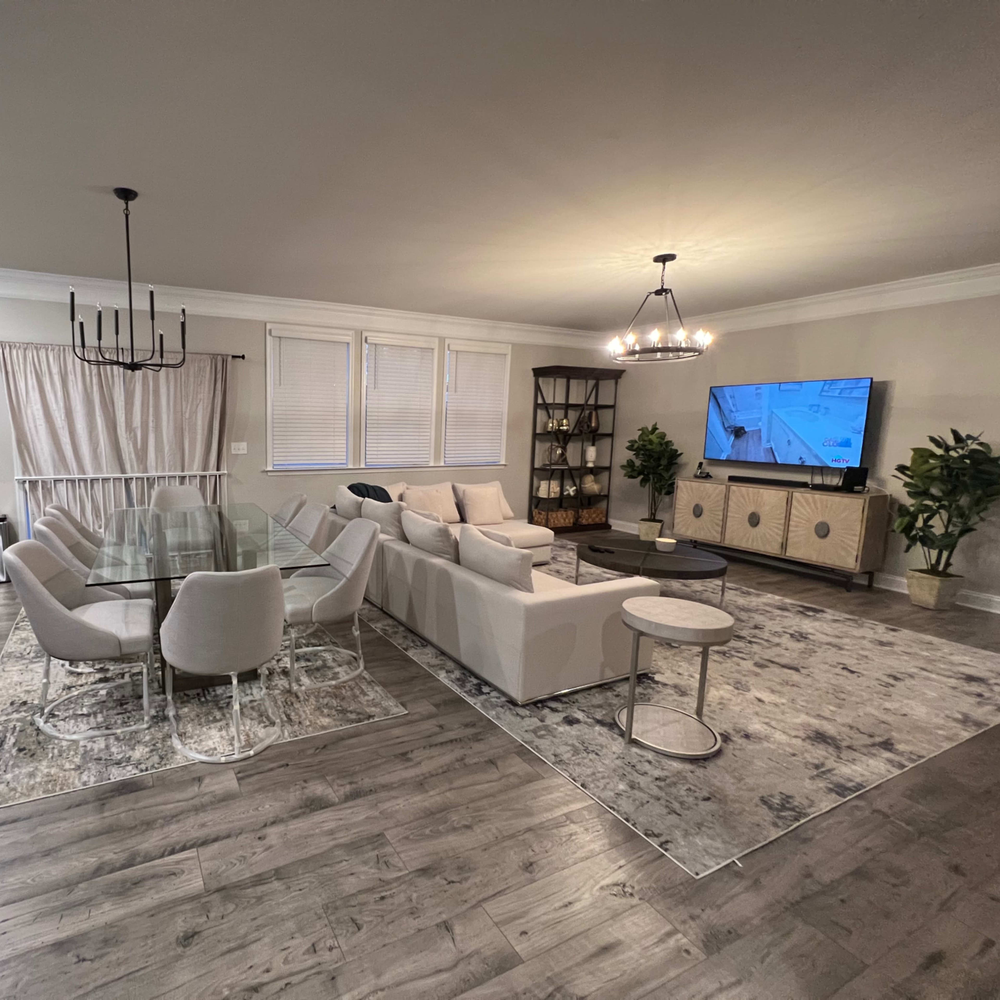 The living room features a glass dining table surrounded by upholstered chairs, a sectional sofa, a TV on a stand, and a decorative bookshelf against the wall.