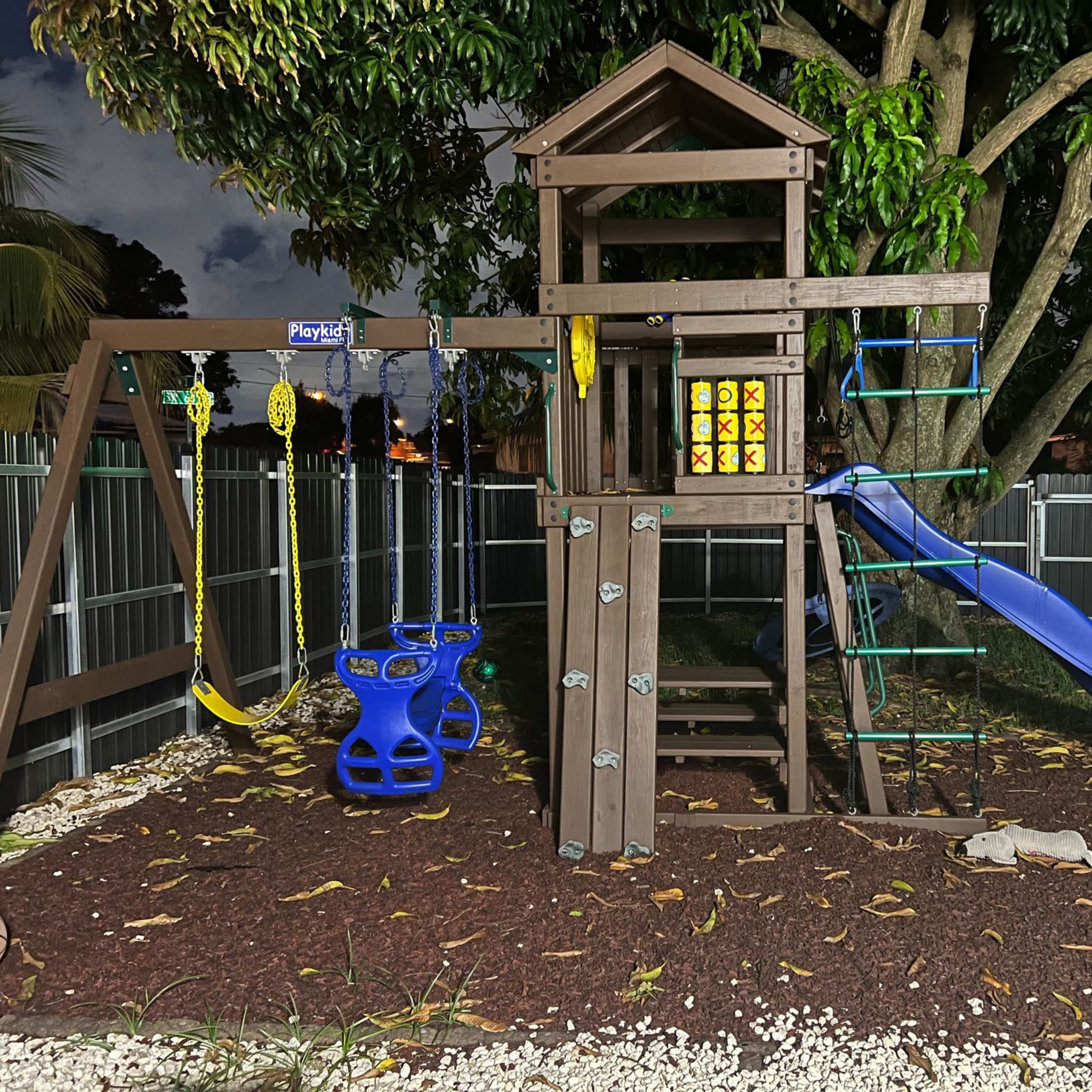 A wooden playground set with a slide, swings, and a climbing structure, situated on grass and surrounded by a fence.