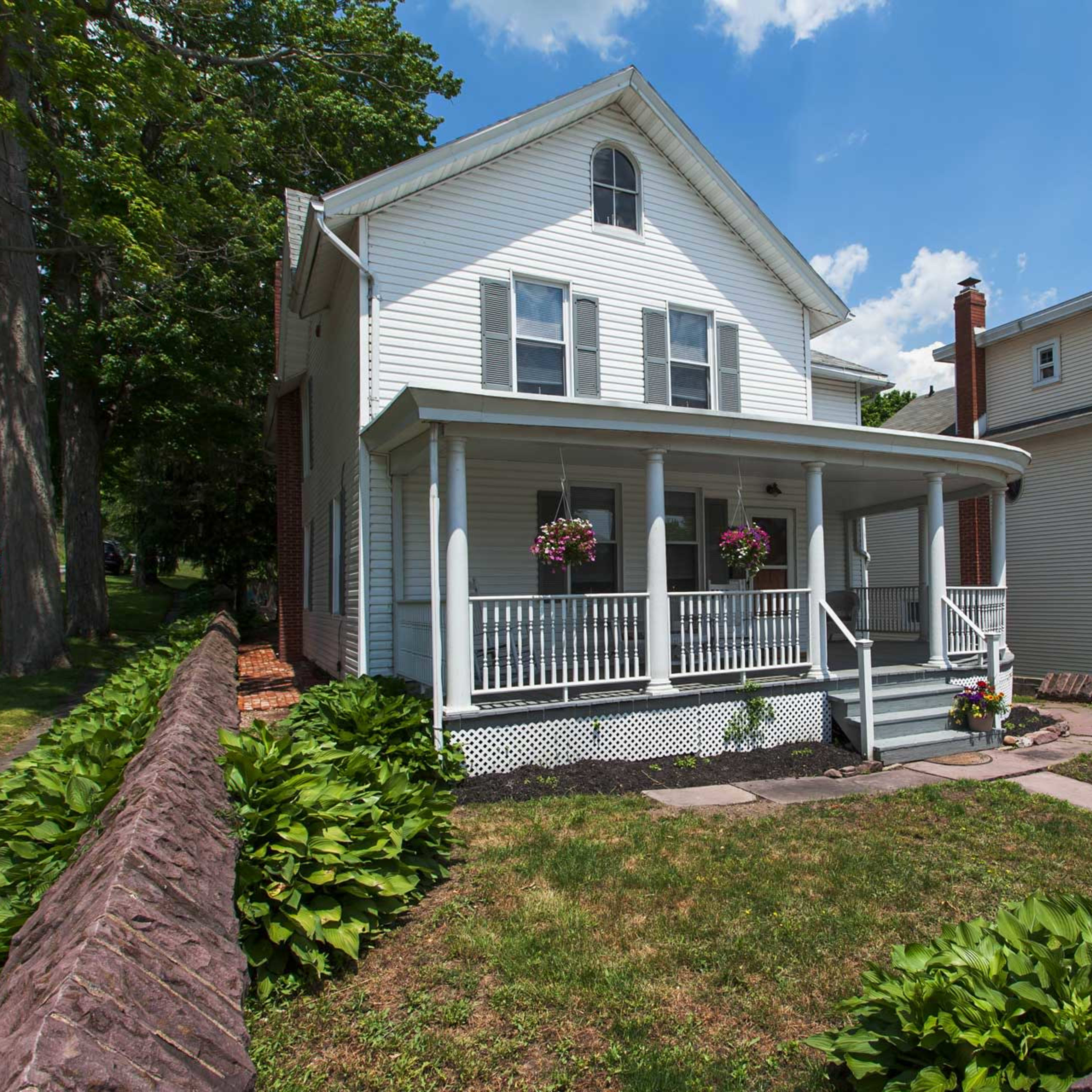 A two-story white house with a front porch, surrounded by a grassy lawn and a low stone wall.