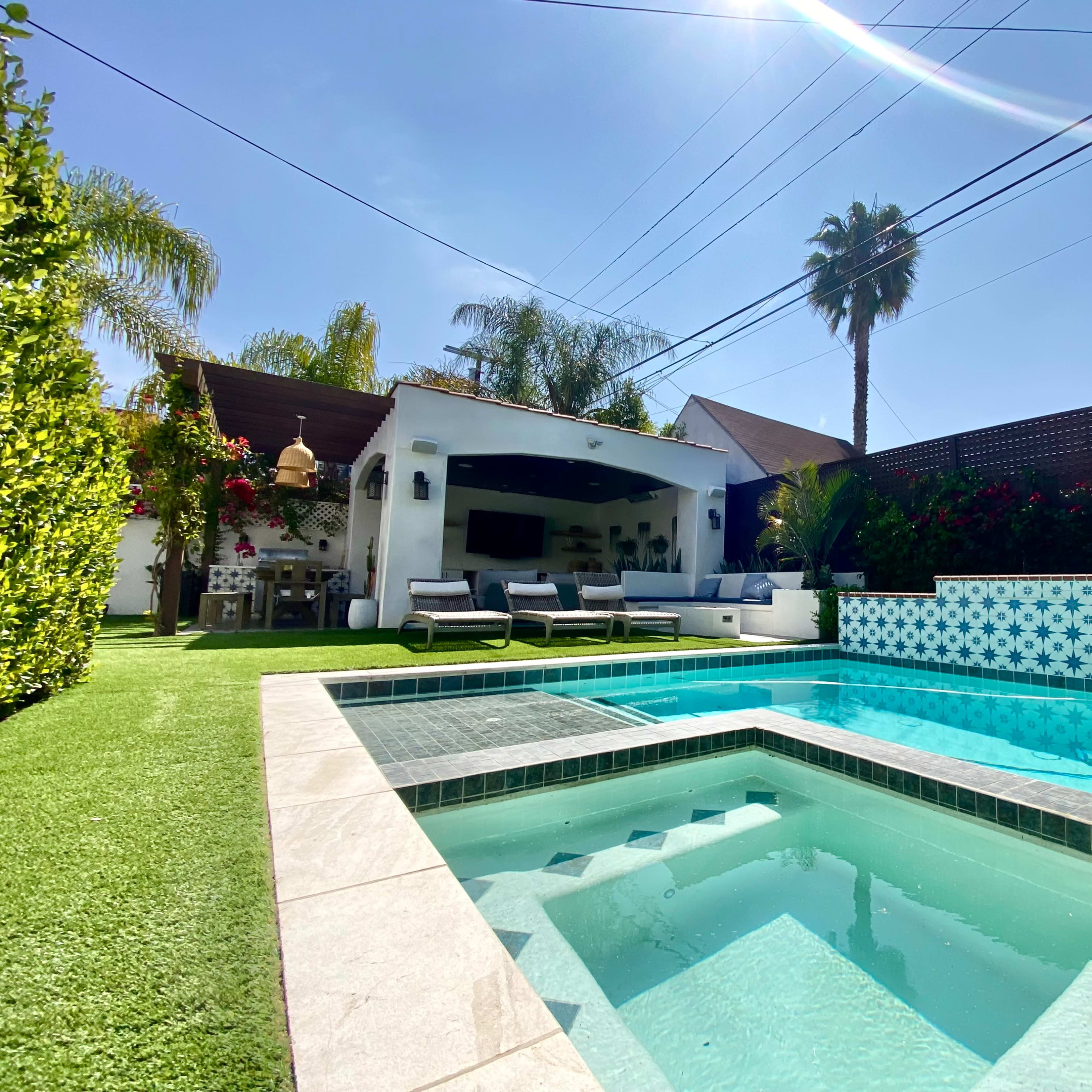 A backyard with a swimming pool featuring tile designs, lounge chairs, and a house partially visible behind it, surrounded by palm trees and greenery.
