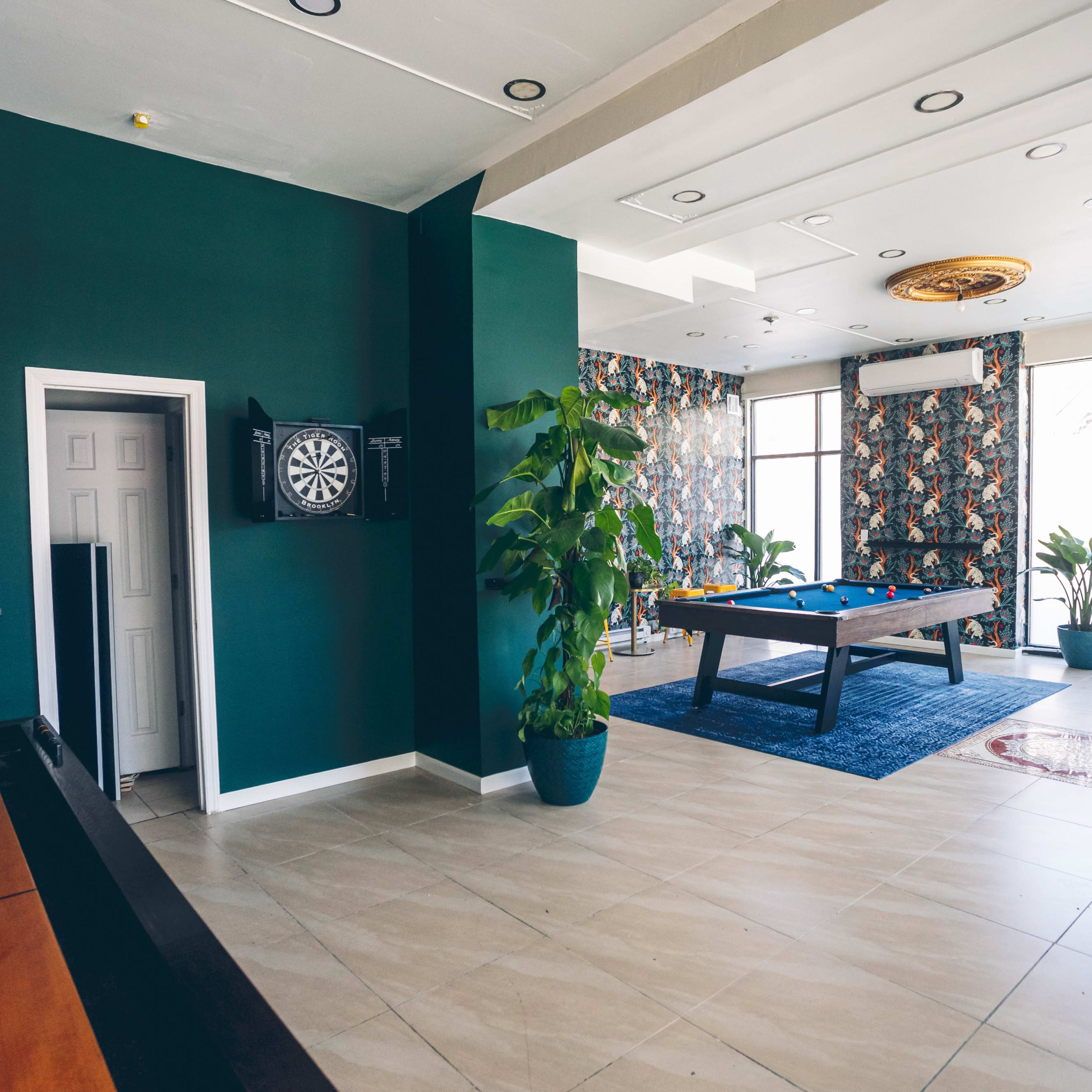 A recreational room with a pool table, a dartboard, and seating surrounded by indoor plants and decorative wallpaper.