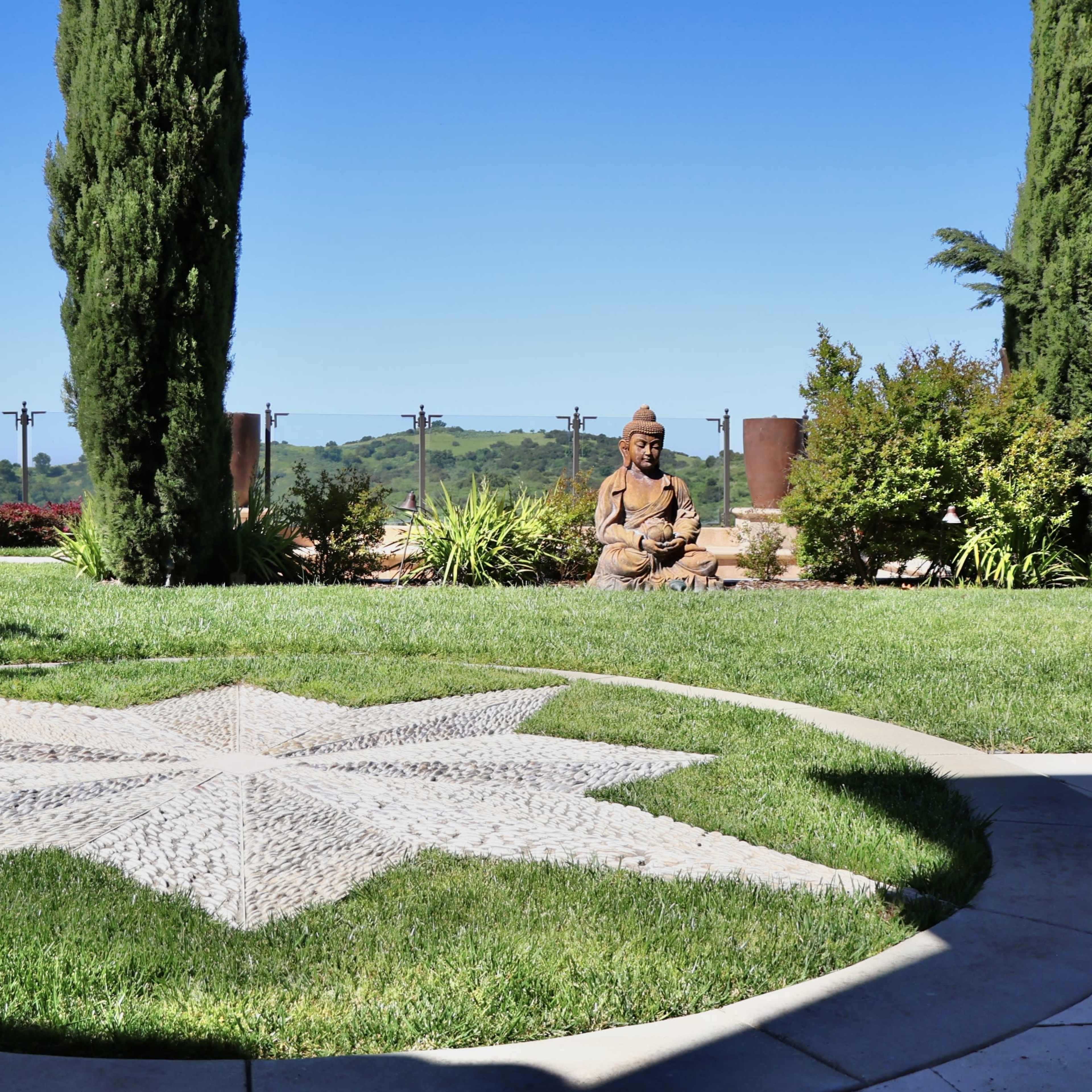 A landscaped garden featuring a stone star pattern embedded in grass, with a seated statue and lush greenery in the background.
