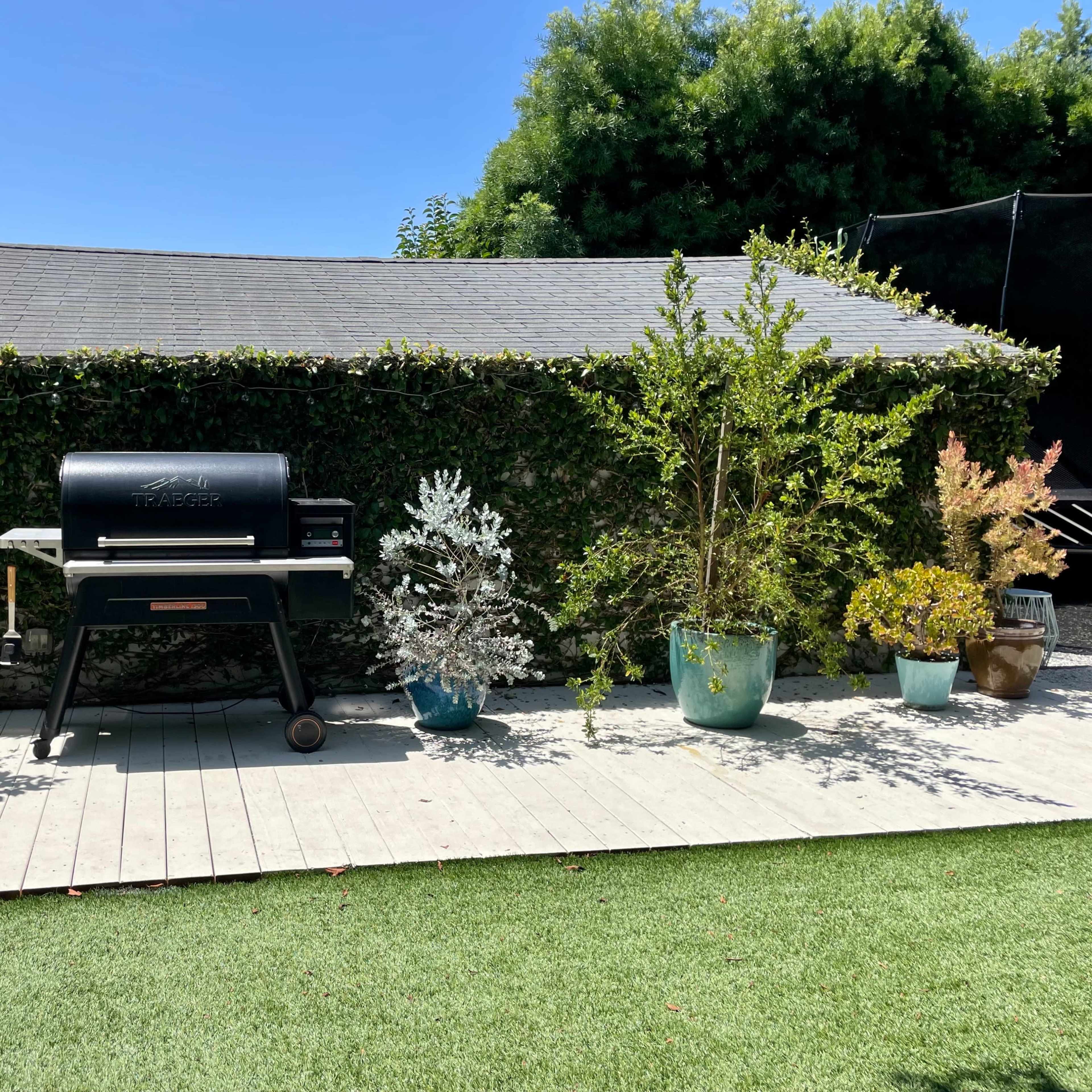 A black grill is positioned on a wooden deck surrounded by various potted plants and a green lawn.