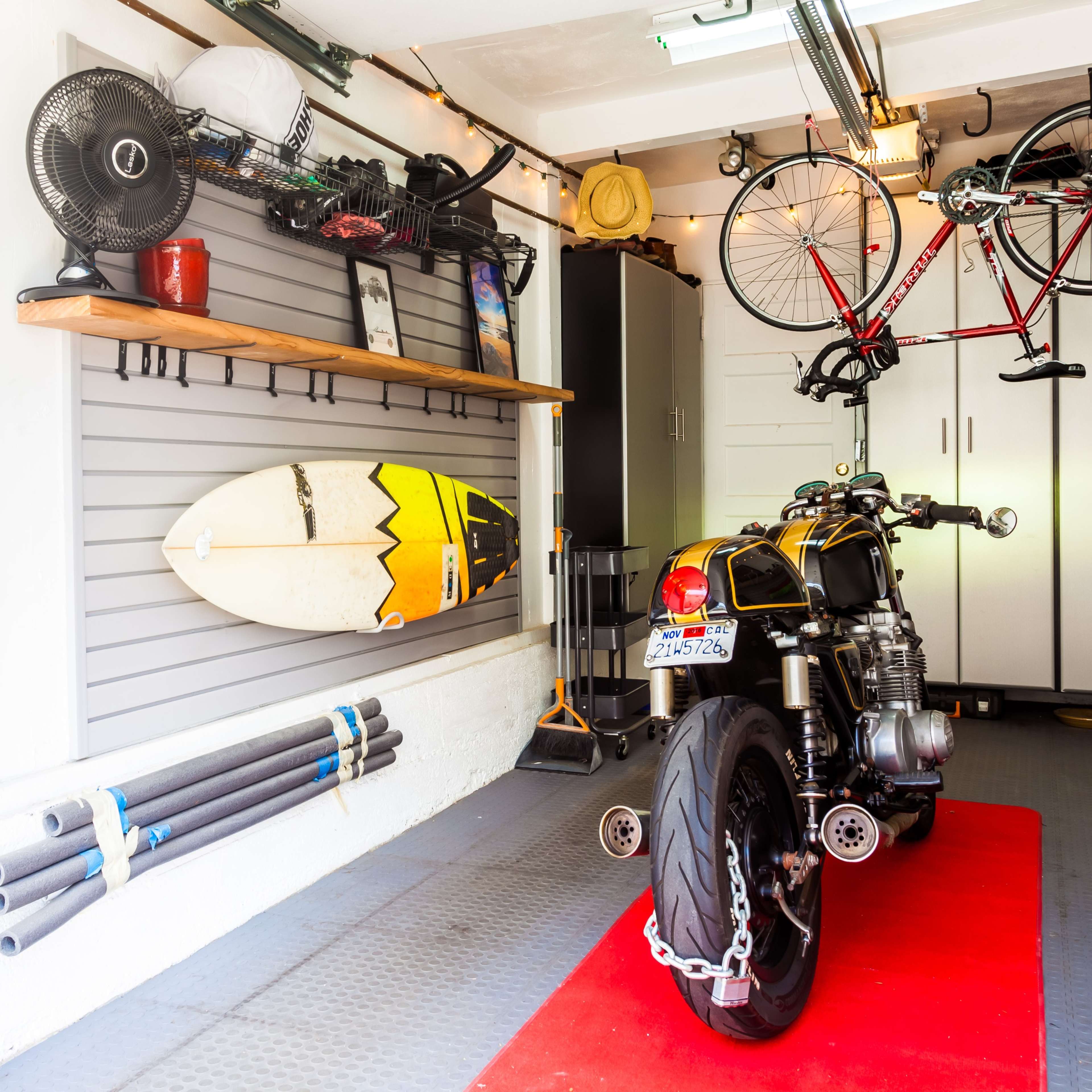 A neatly organized garage with a motorcycle on a red mat, a surfboard leaning against the wall, a bicycle hanging from the ceiling, and storage cabinets filled with various tools and equipment.