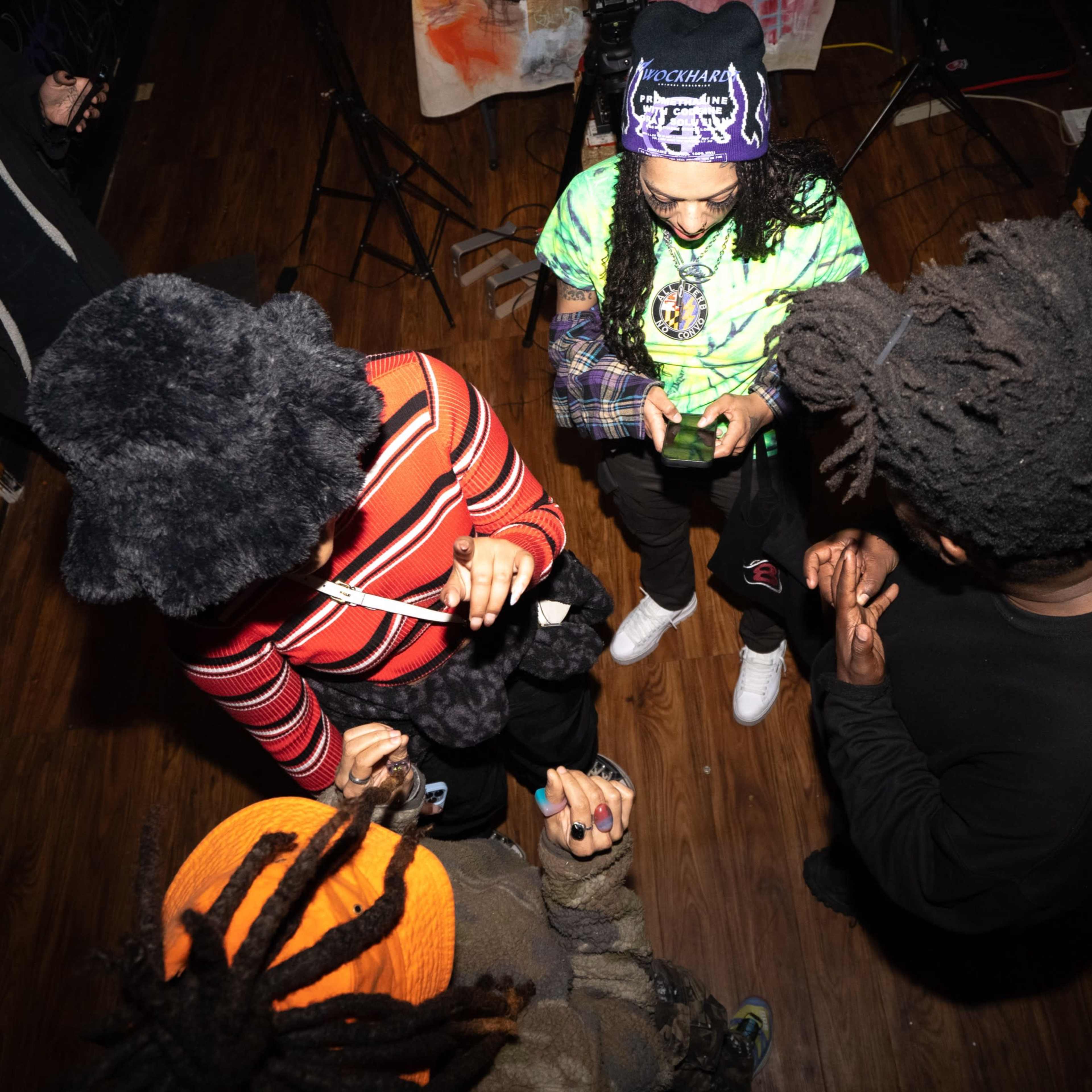 A group of four people are gathered in a dimly lit room, engaged in using their phones while wearing various styles of clothing and headwear.