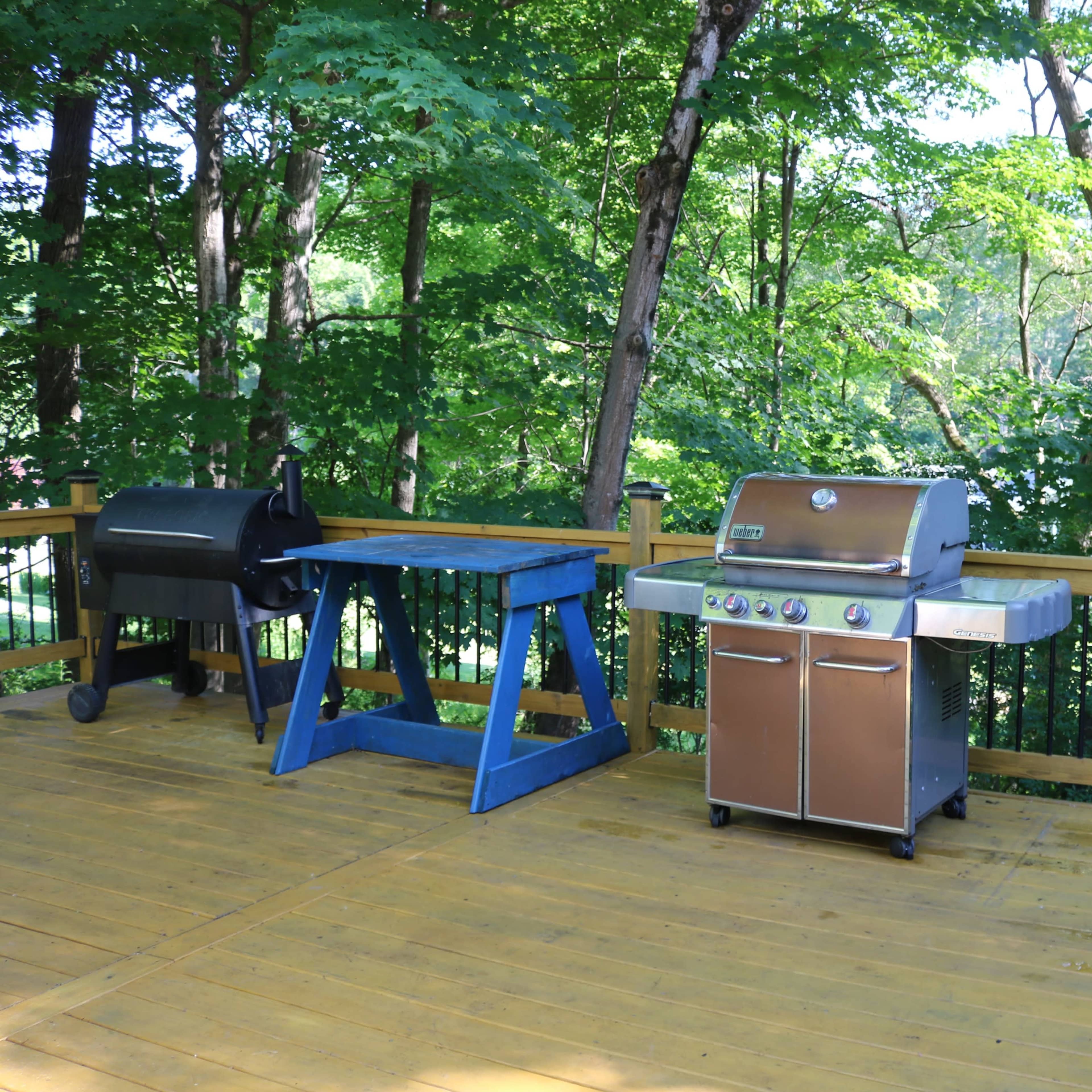 A wooden deck with a blue work table and a stainless steel grill, surrounded by lush green trees.