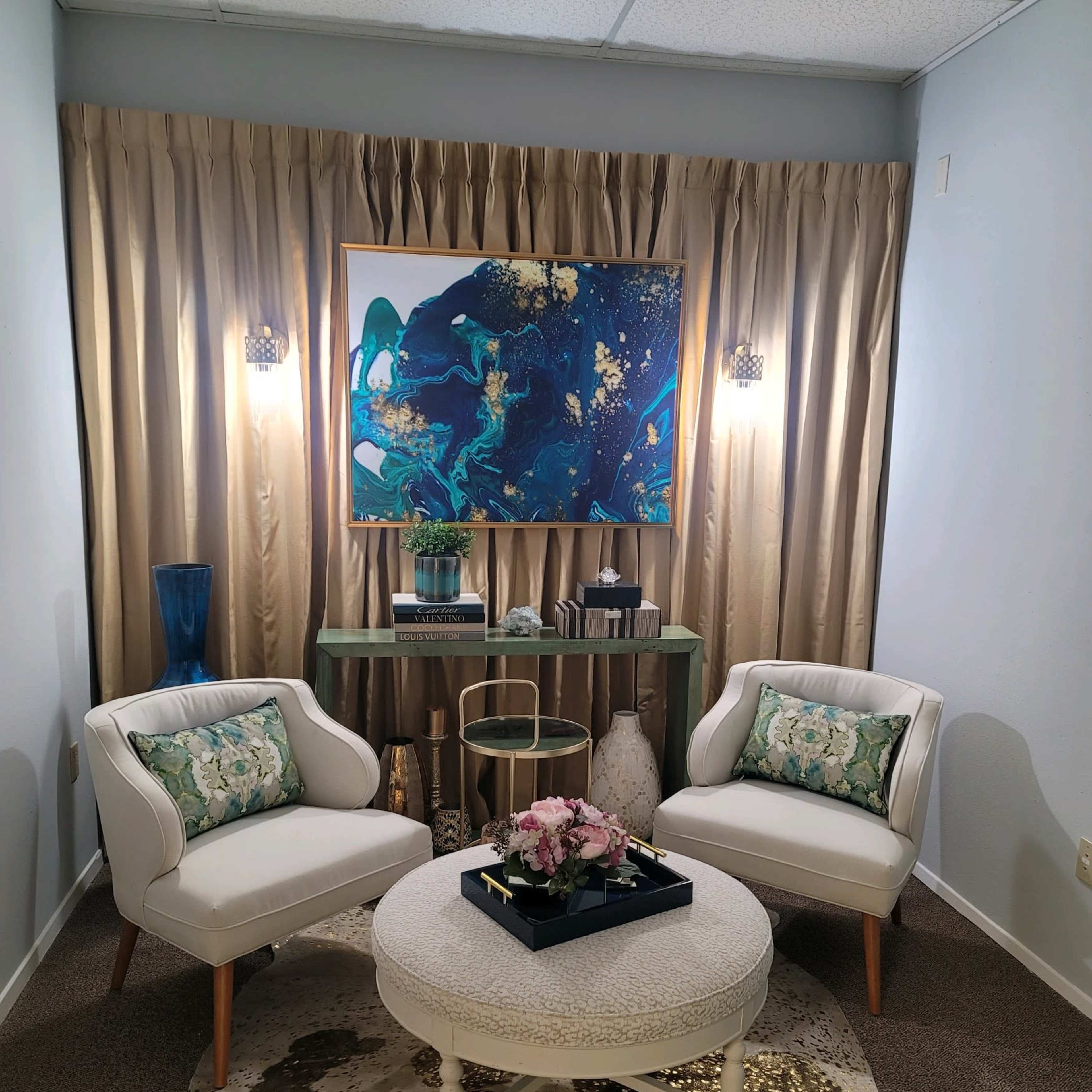 A cozy seating area with two white chairs, a round coffee table adorned with flowers, and a decorative backdrop consisting of a large painting and elegant curtains.
