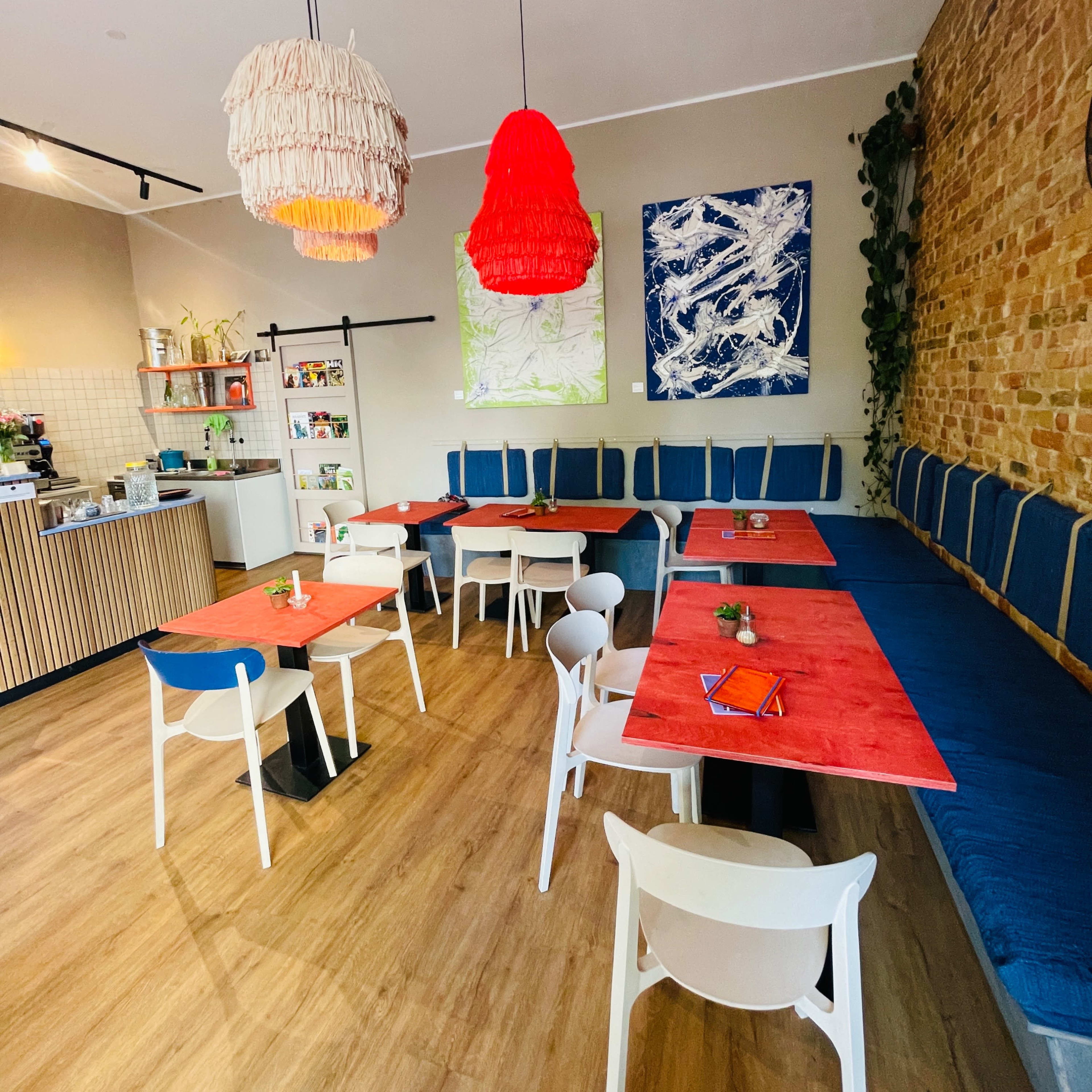 The interior of a café features wooden flooring, brightly colored tables, and a mix of seating along a textured brick wall.