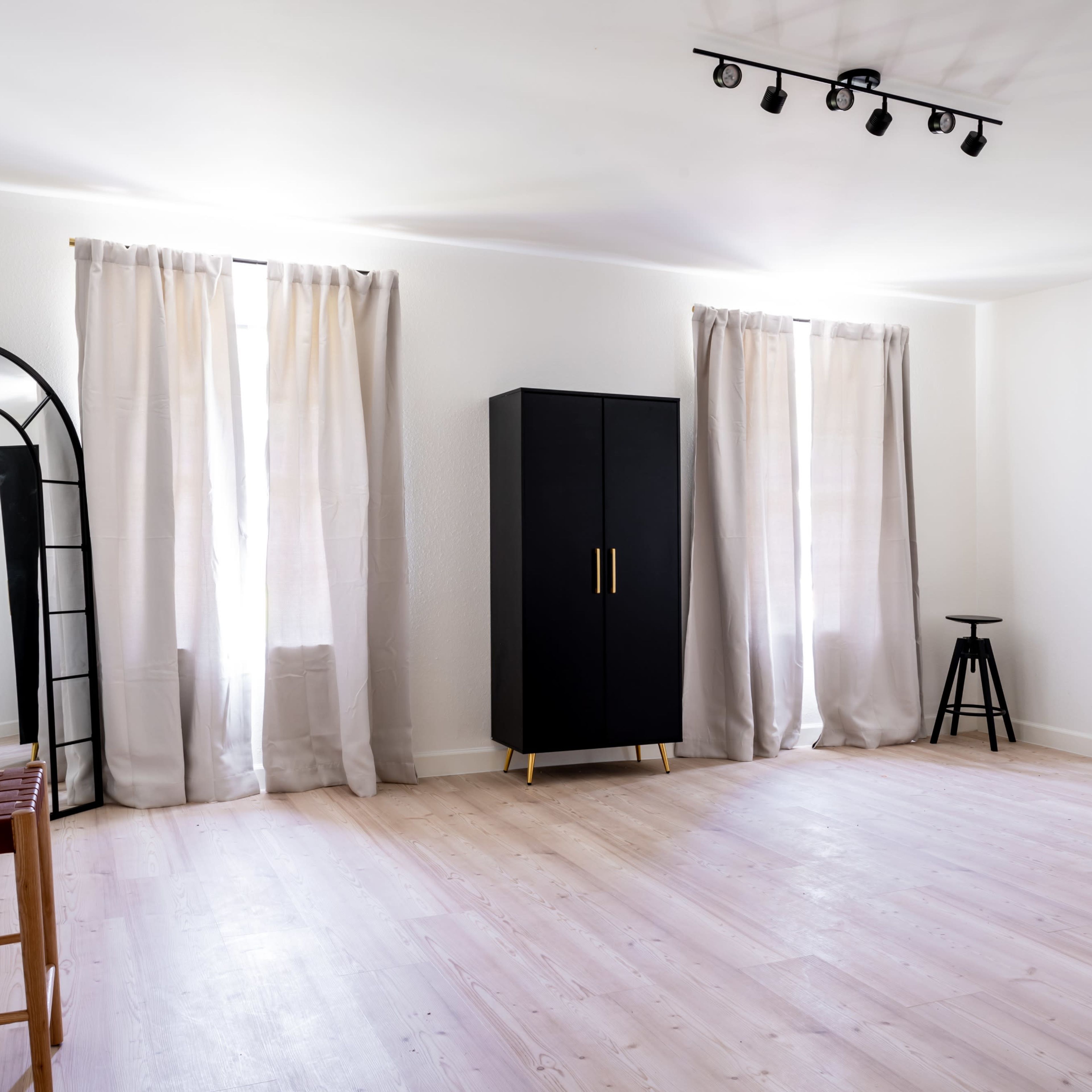 The room features two windows with curtains, a tall black cabinet, a stool, a full-length mirror, and a wooden chair on a light-colored floor.