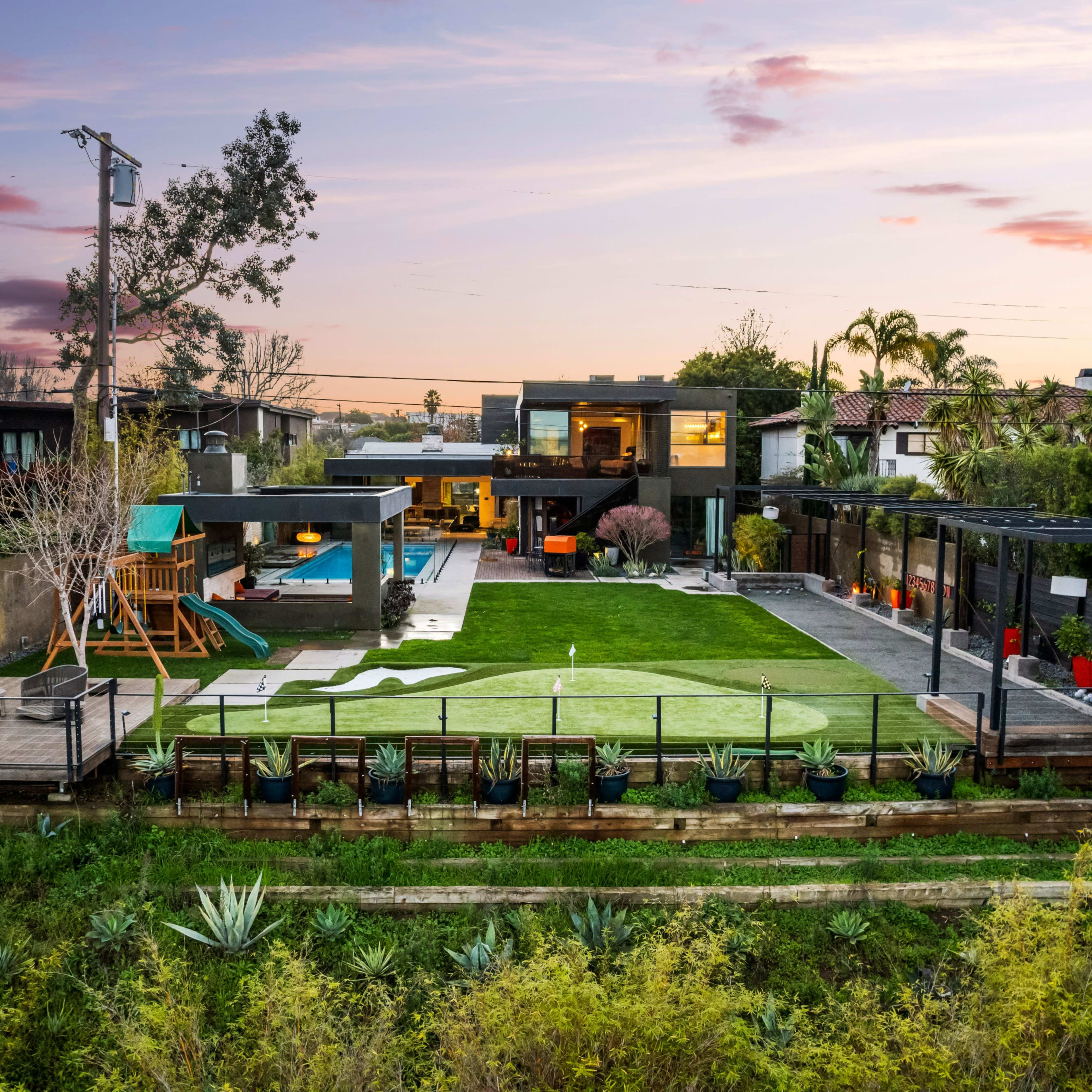 A modern house with a landscaped yard, swimming pool, and a playground is shown against a twilight sky.