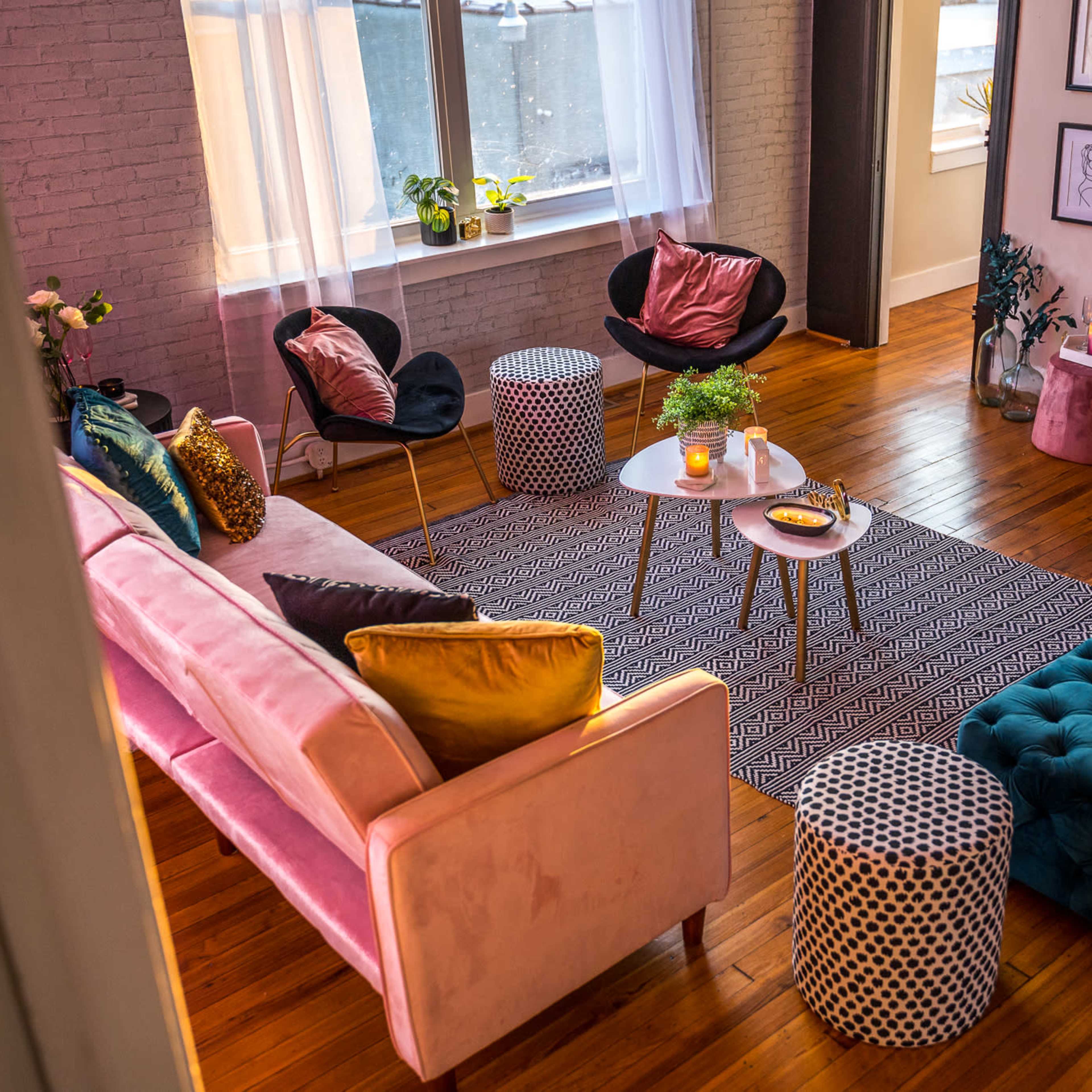 A cozy living room features a mix of seating options, including a pink sofa, black chairs, and various accent tables arranged around a patterned area rug.