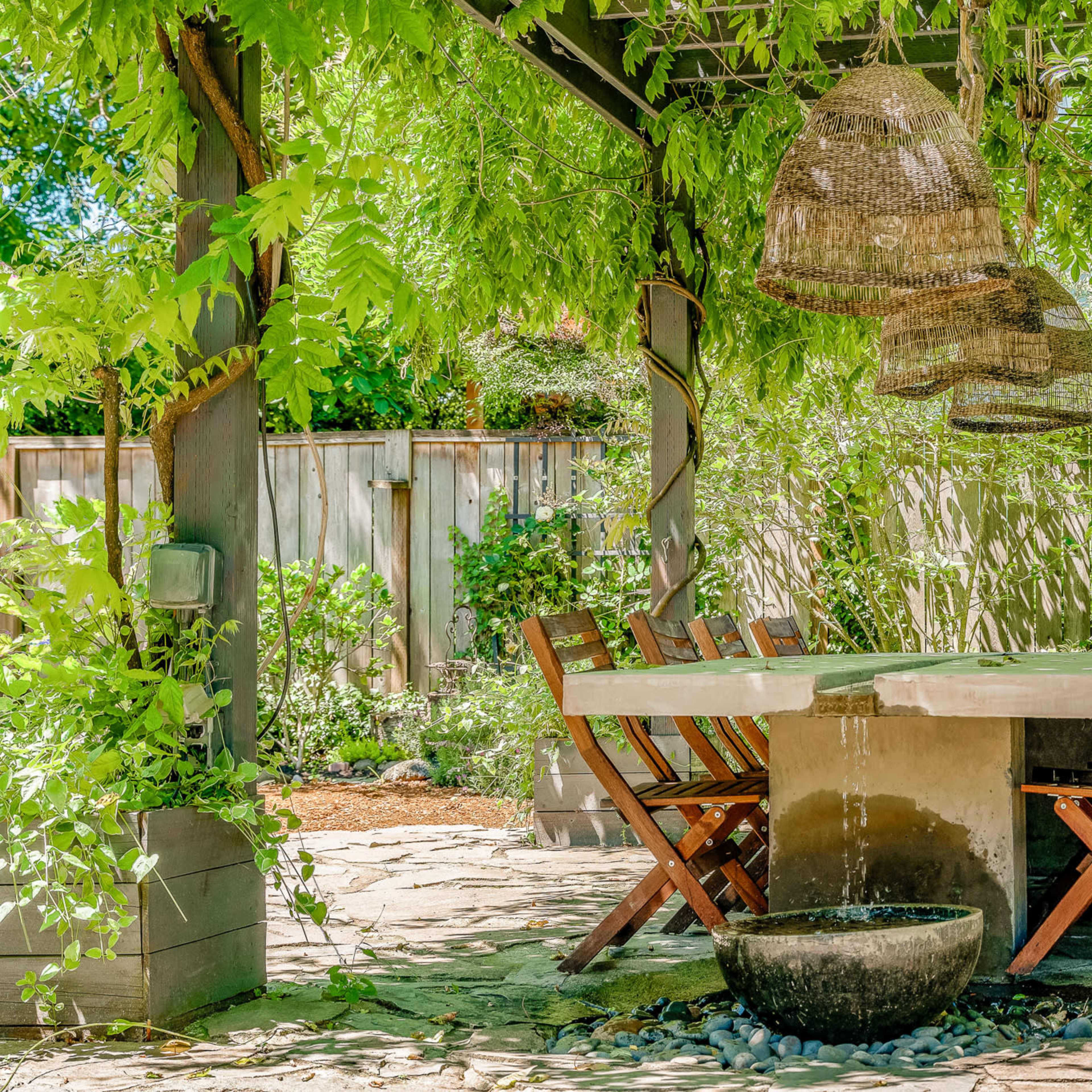 A wooden dining table surrounded by chairs sits under a pergola adorned with hanging lanterns and greenery.