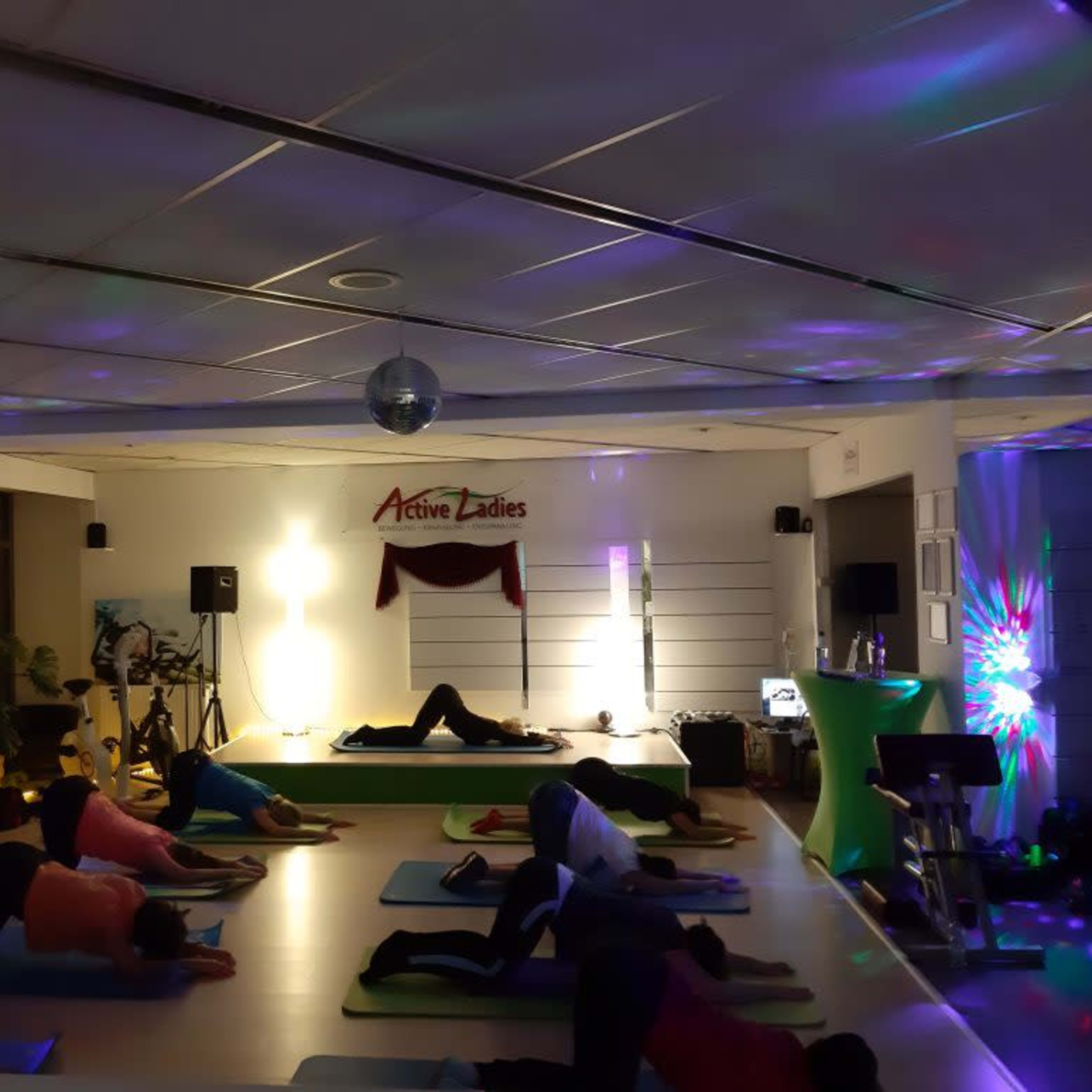 A yoga class takes place in a dimly lit studio with participants lying on mats while colorful lights illuminate the space.