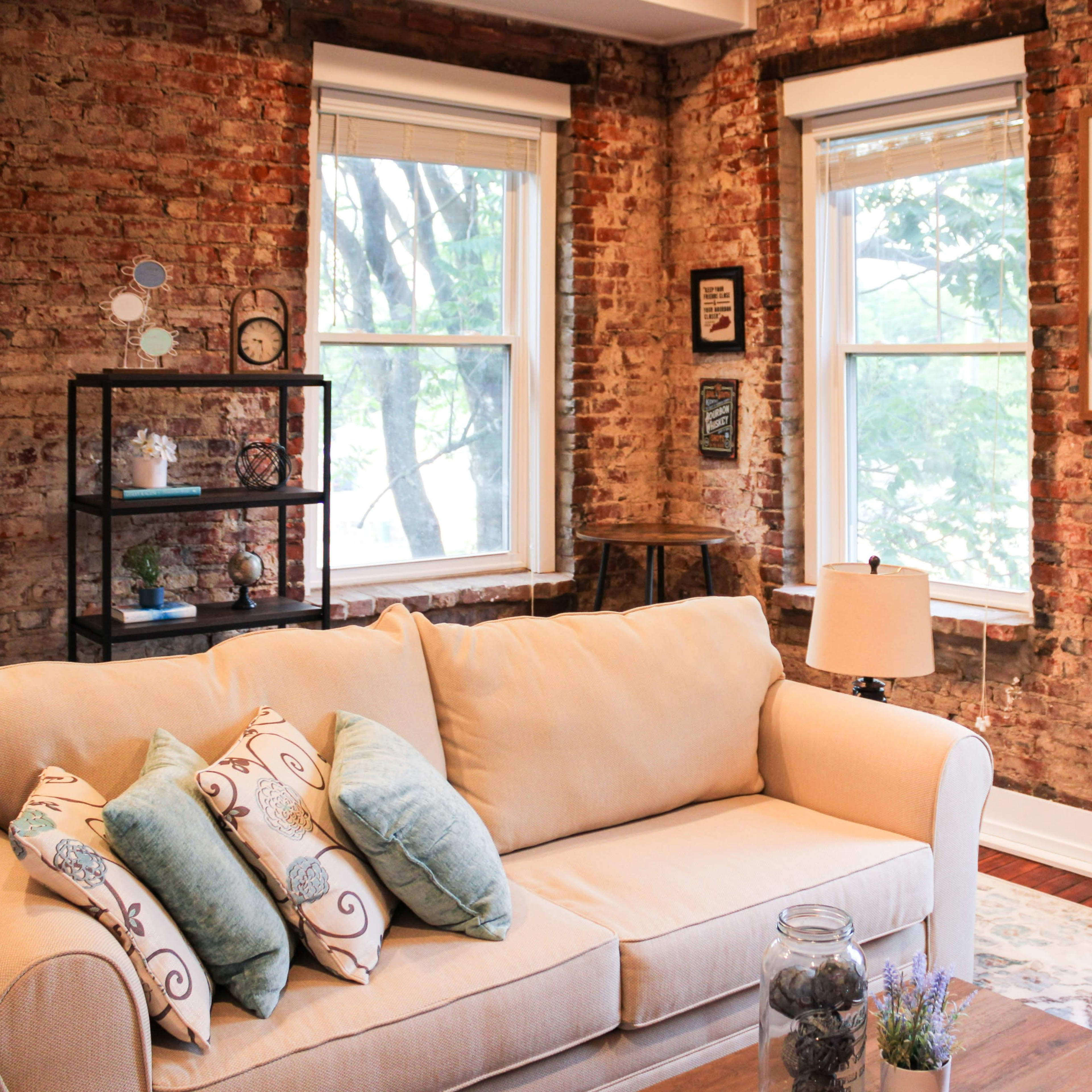 A cozy living room with a beige sofa adorned with decorative pillows, set against a backdrop of exposed brick walls and large windows.