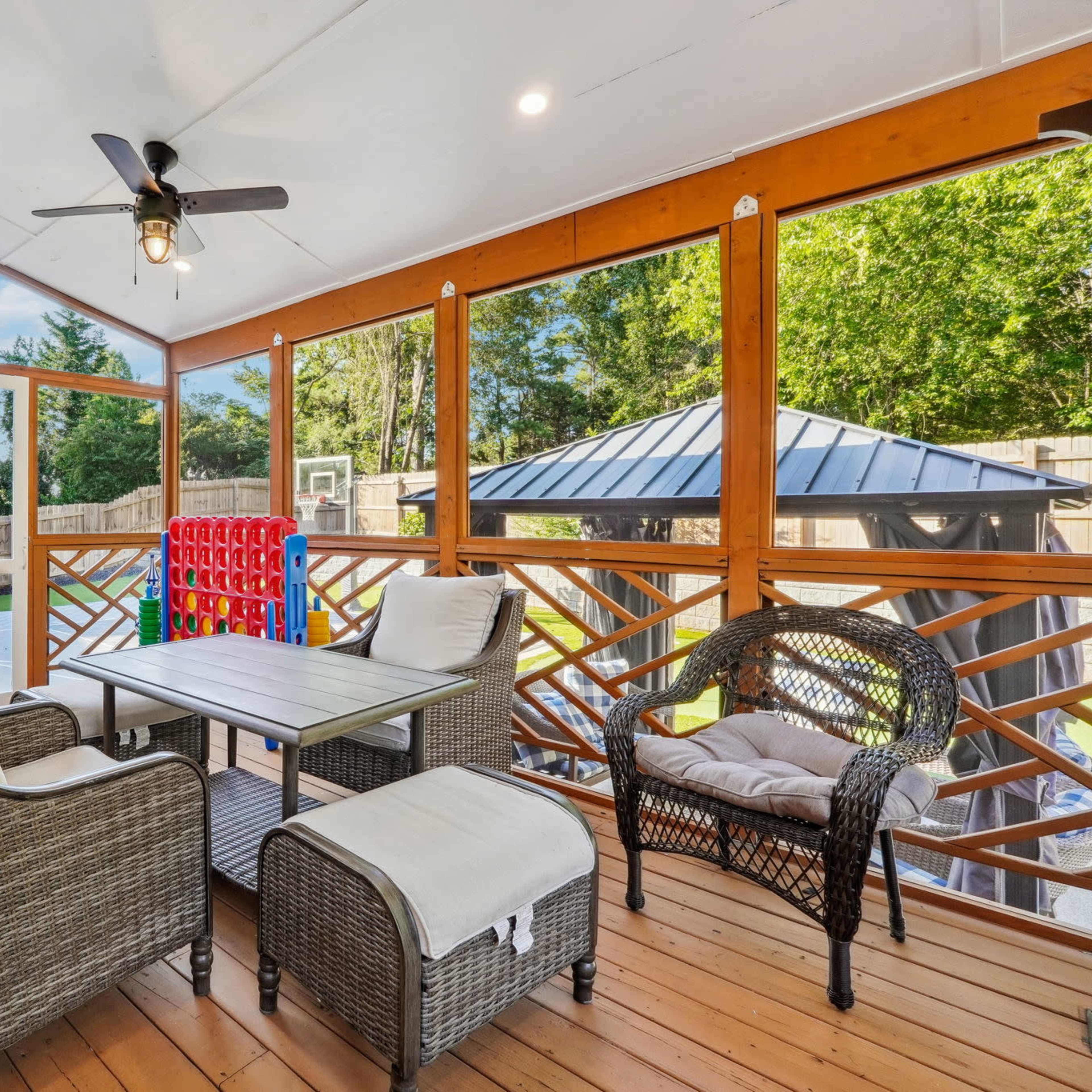 A screened porch with wicker furniture, a ceiling fan, and a view of a backyard featuring a basketball court and a gazebo.