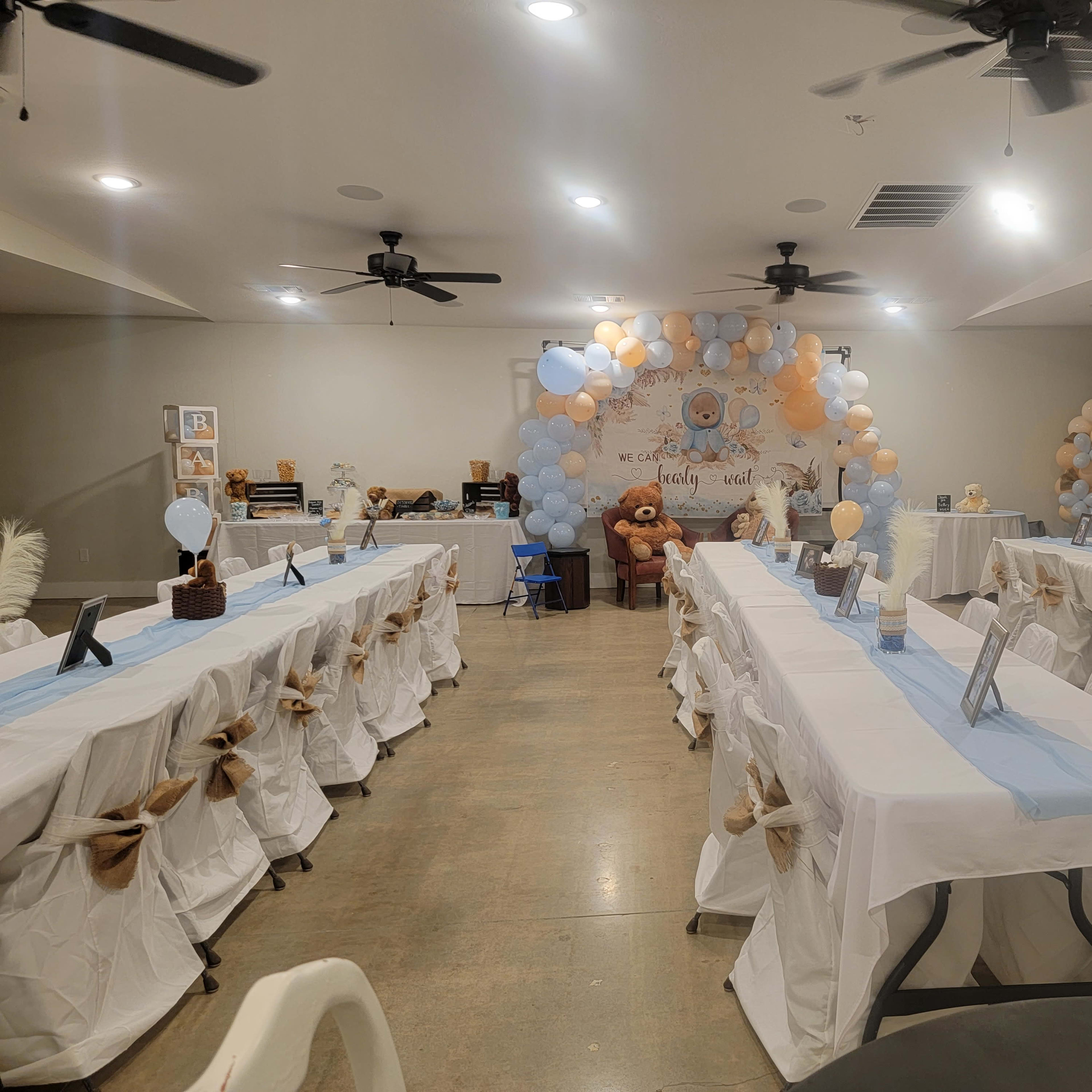 A banquet hall decorated for a celebration, featuring long tables draped with white tablecloths, blue accents, and a backdrop of balloons and stuffed animals.