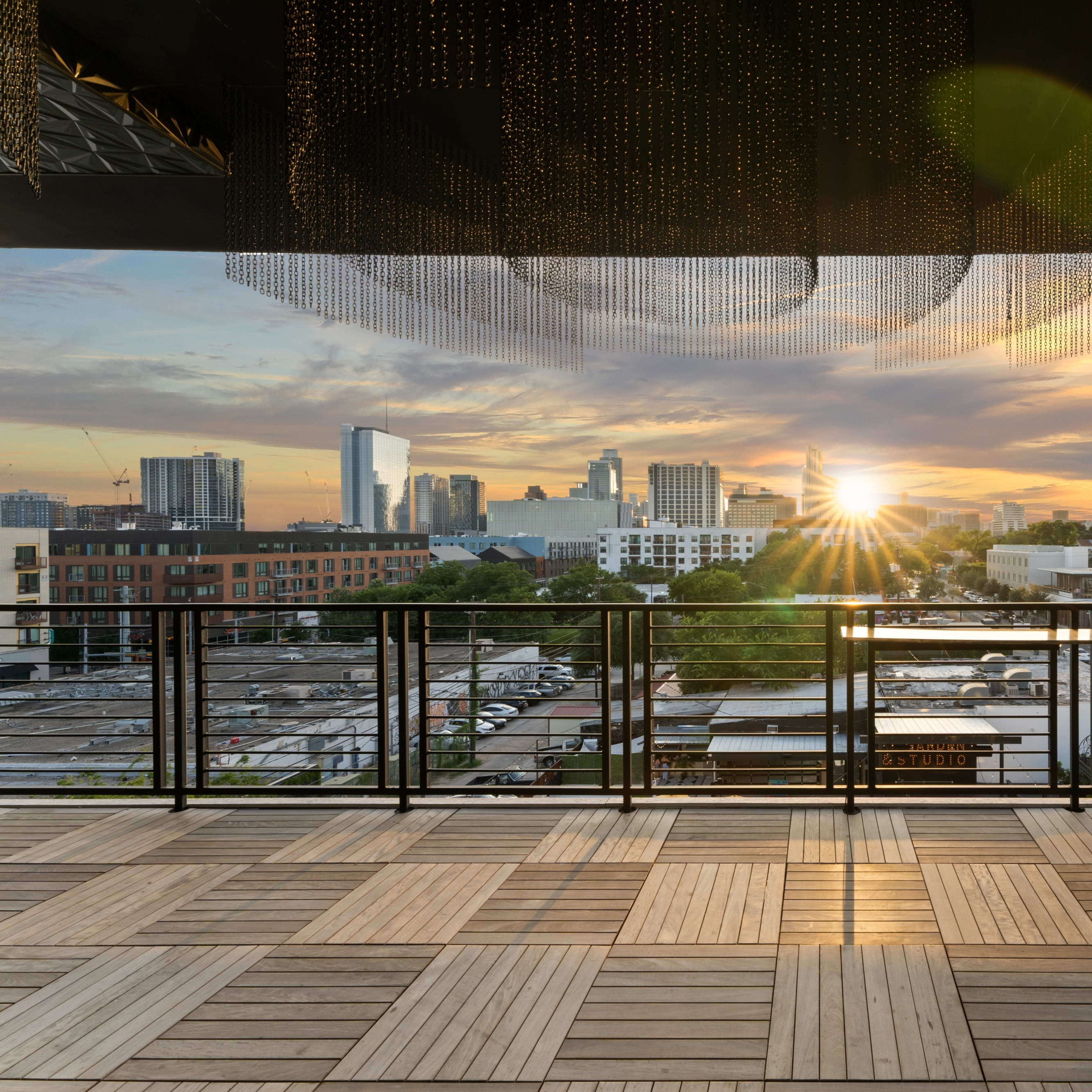 A rooftop terrace with wooden flooring overlooking a city skyline at sunset, featuring buildings and a radiant sun in the background.