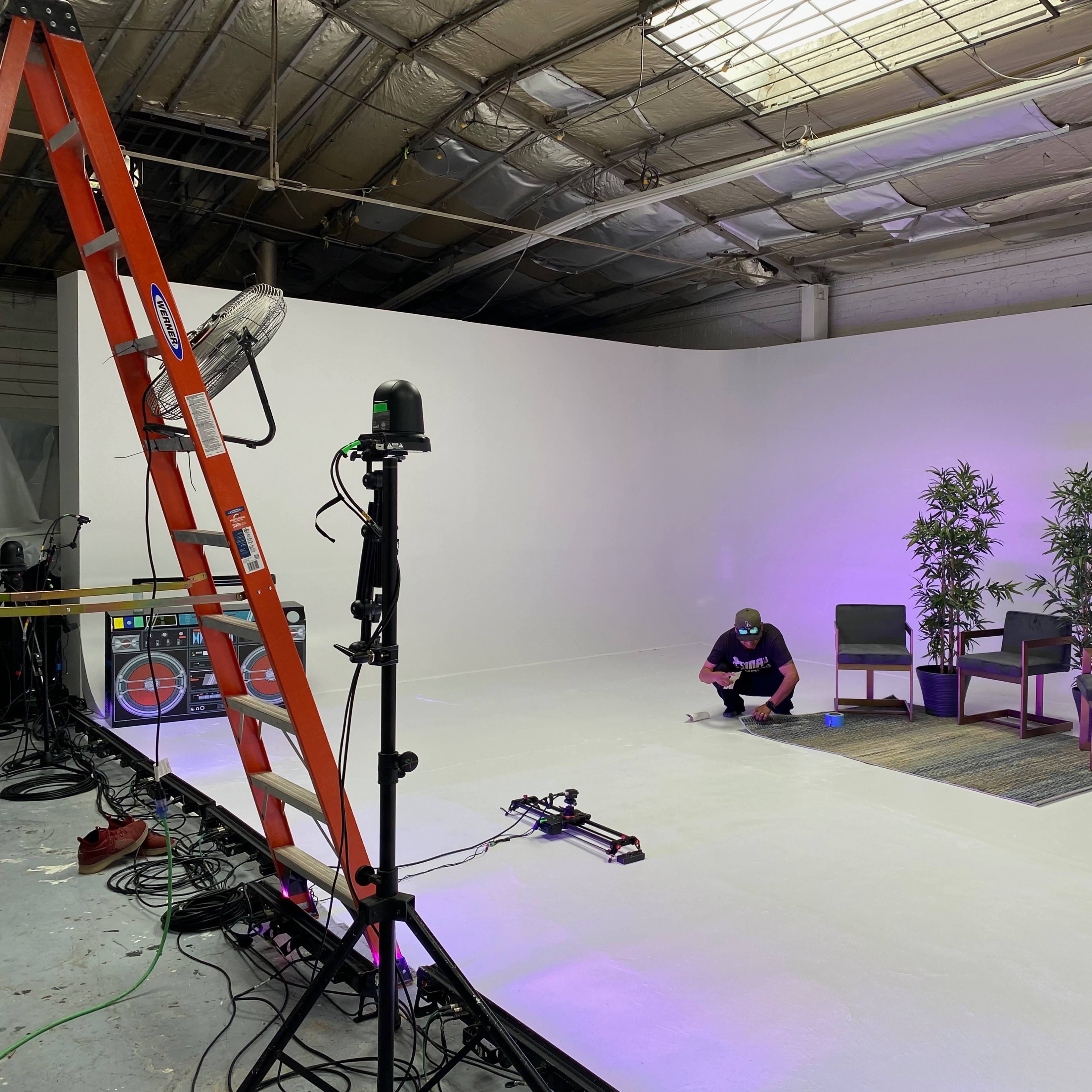 A person kneels on a white floor in an empty studio space, surrounded by a ladder, lighting equipment, and a small seating area with potted plants.