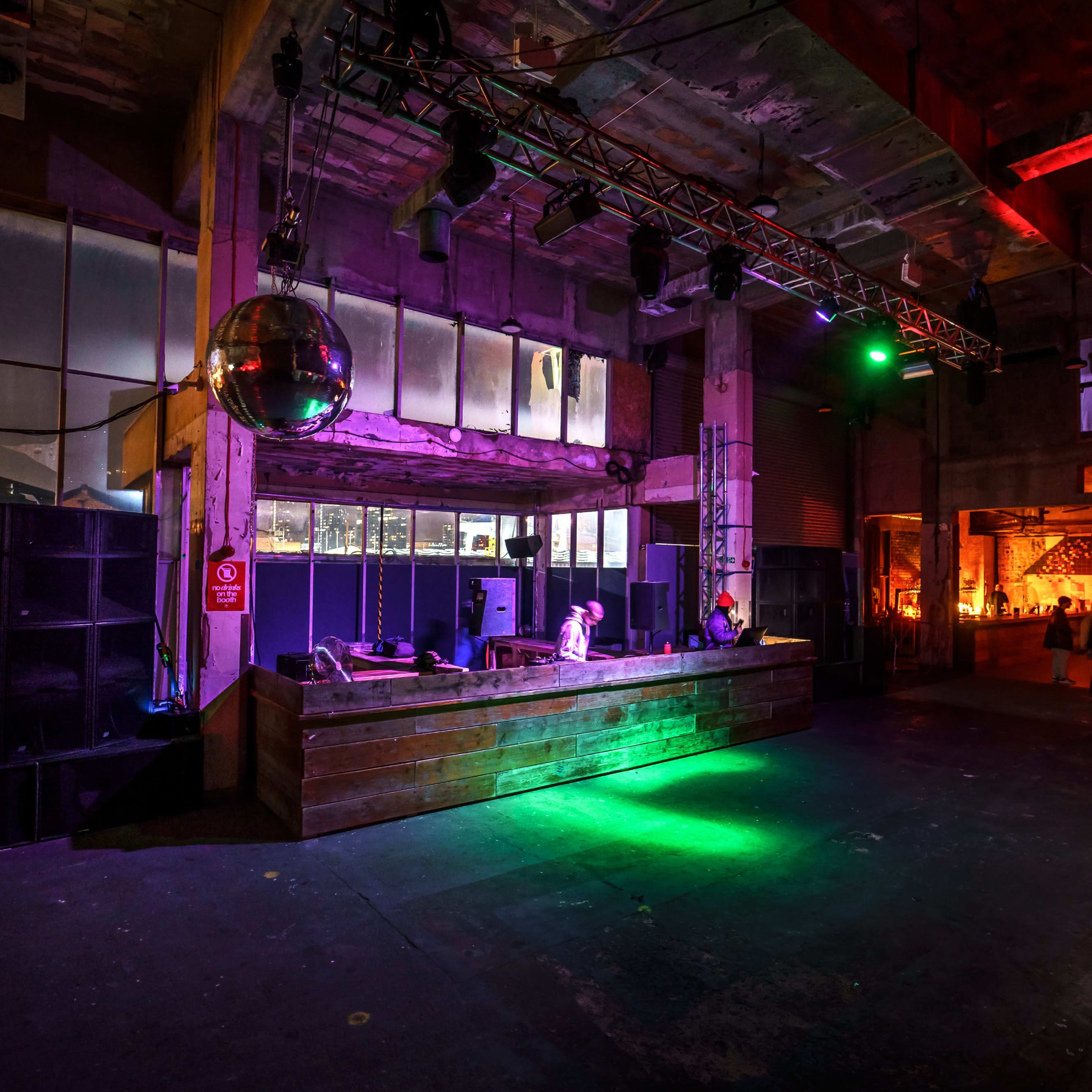 A DJ booth with a disco ball is set up in a spacious, dimly lit venue, with colorful lights illuminating the area.