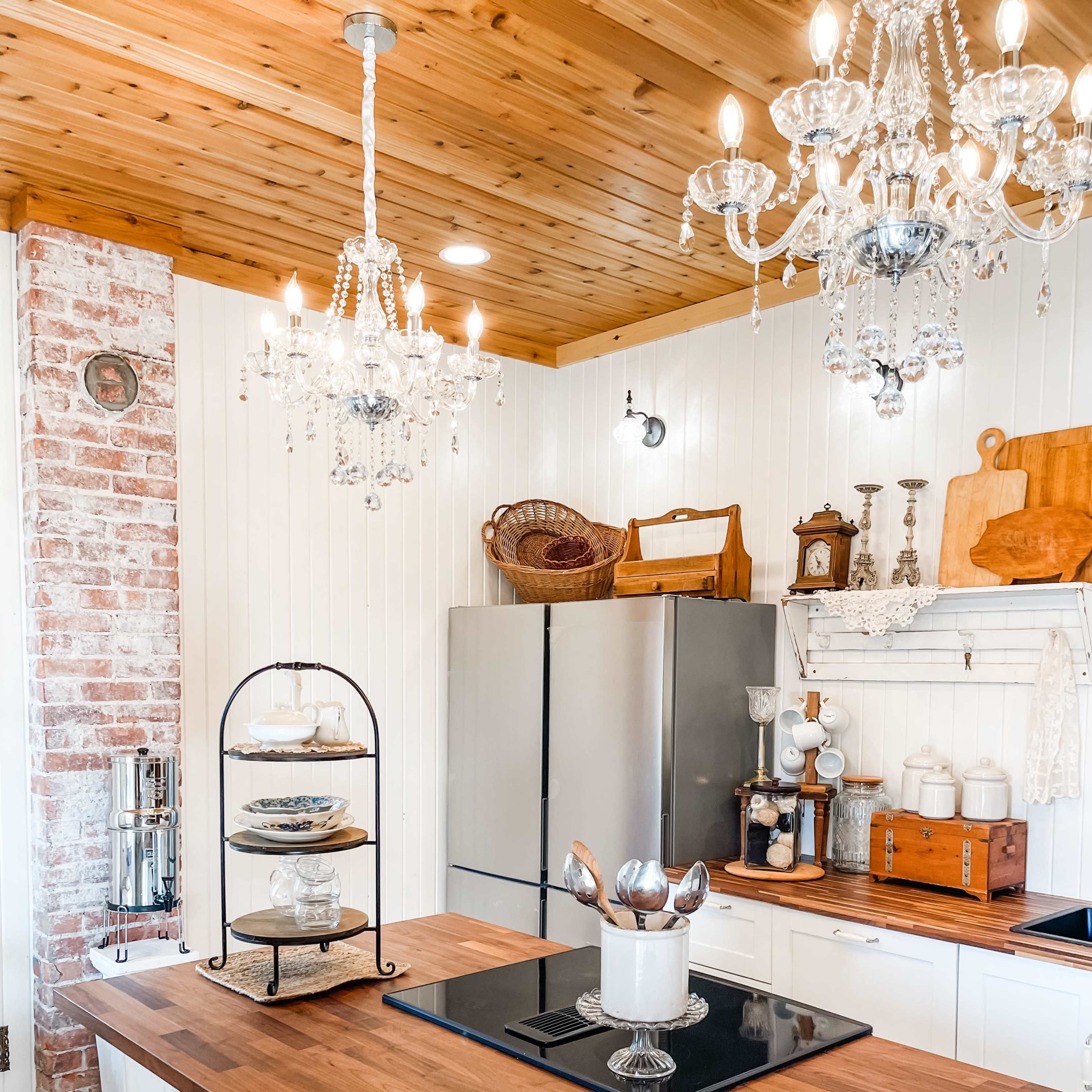 A modern kitchen features a wooden island with a stand holding dishes, stainless steel appliances, and two ornate chandeliers hanging from the ceiling.
