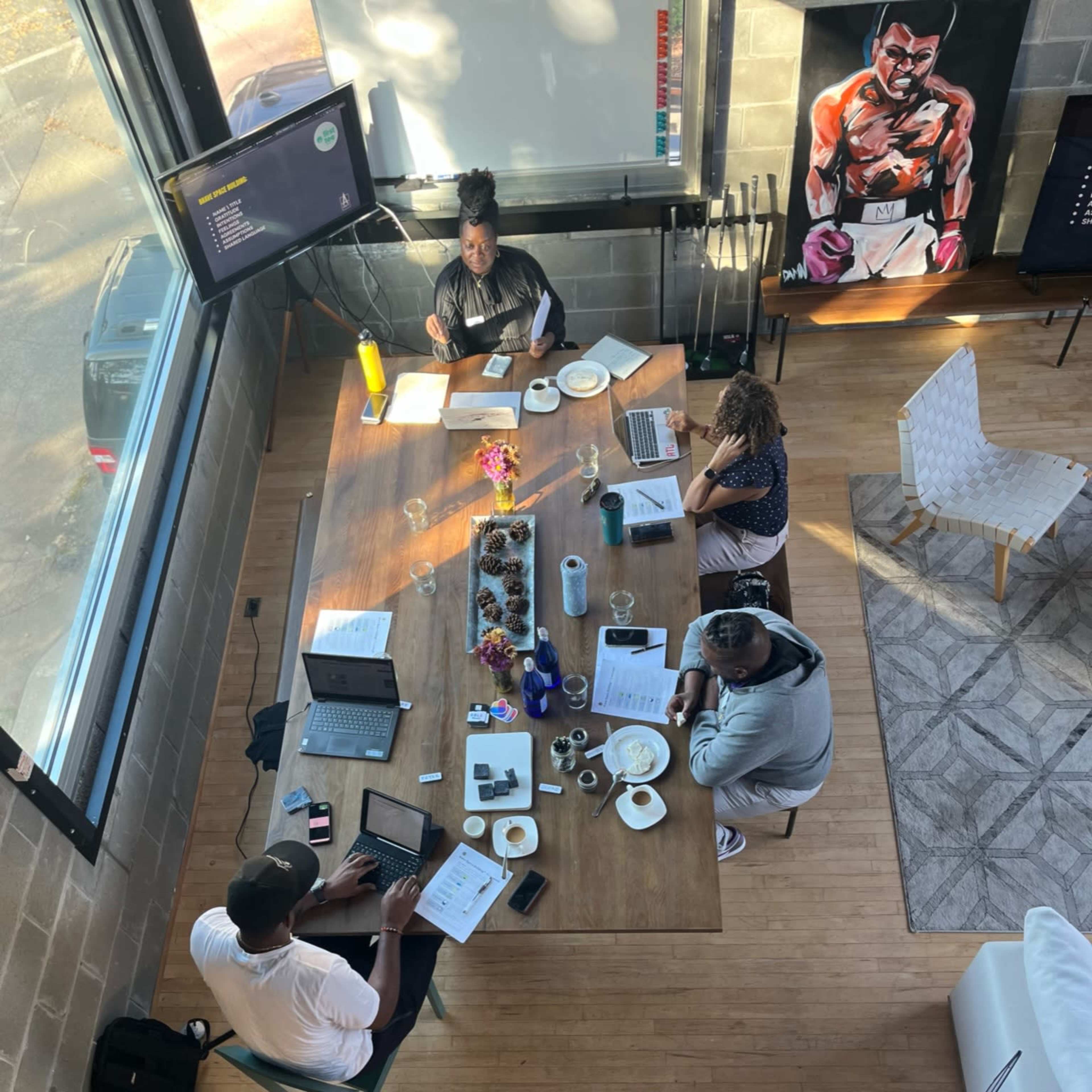A group of four people work collaboratively around a large wooden table filled with laptops and documents in a bright room with large windows.