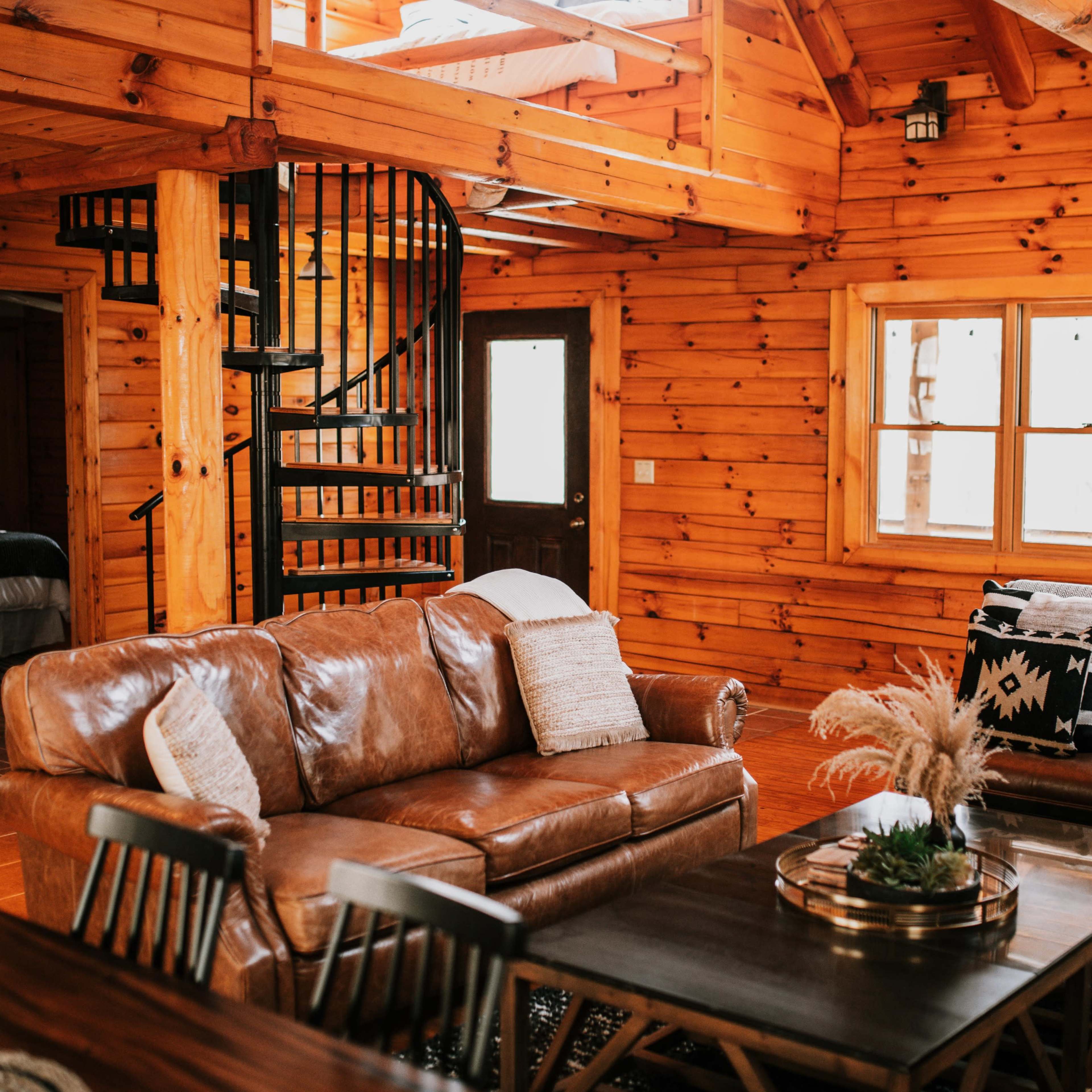 The interior of a log cabin features a cozy living area with a leather sofa, a wooden table, and a spiral staircase leading to a loft.