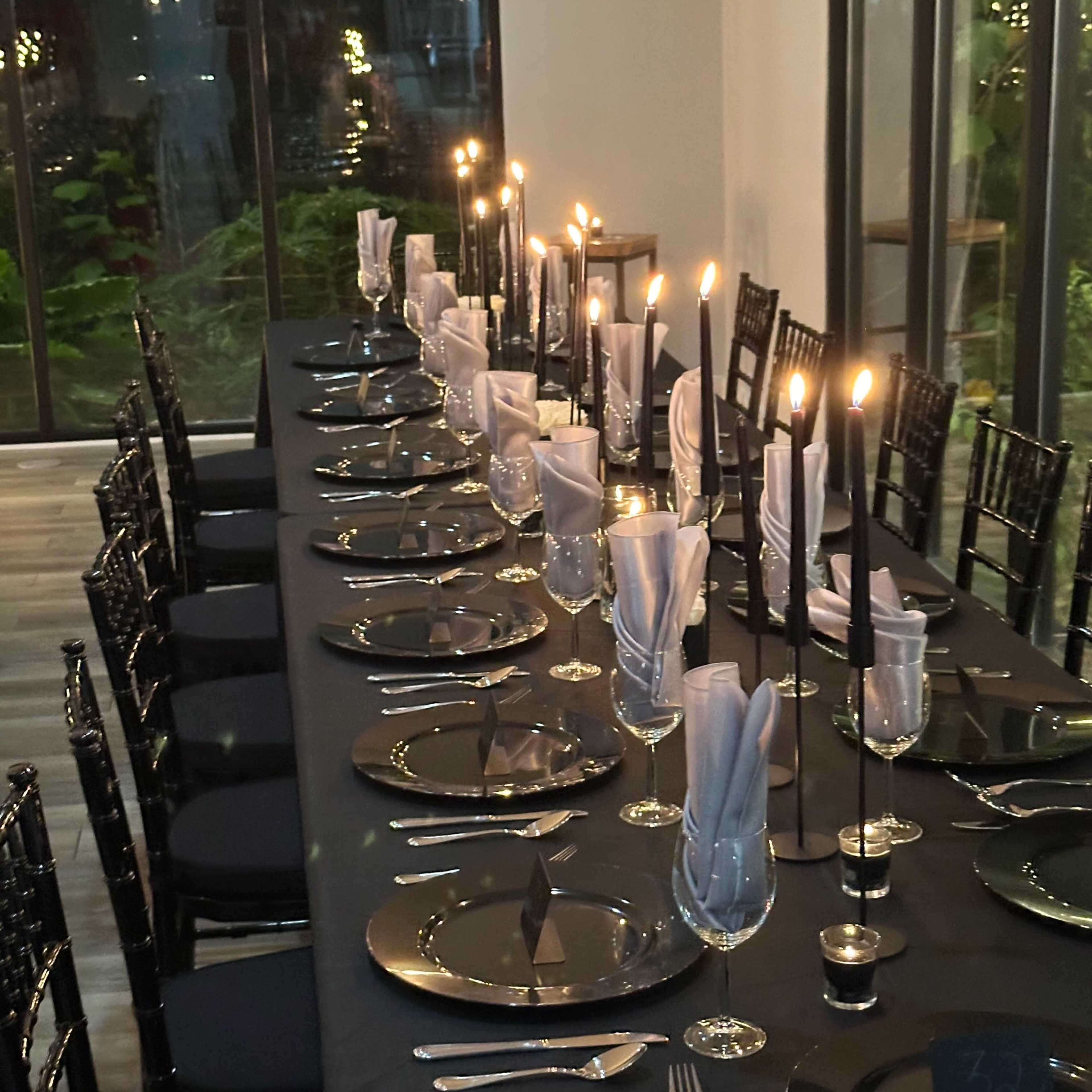 A long dining table is set with black tablecloths, elegant glassware, and lit candles along the center, surrounded by black chairs.