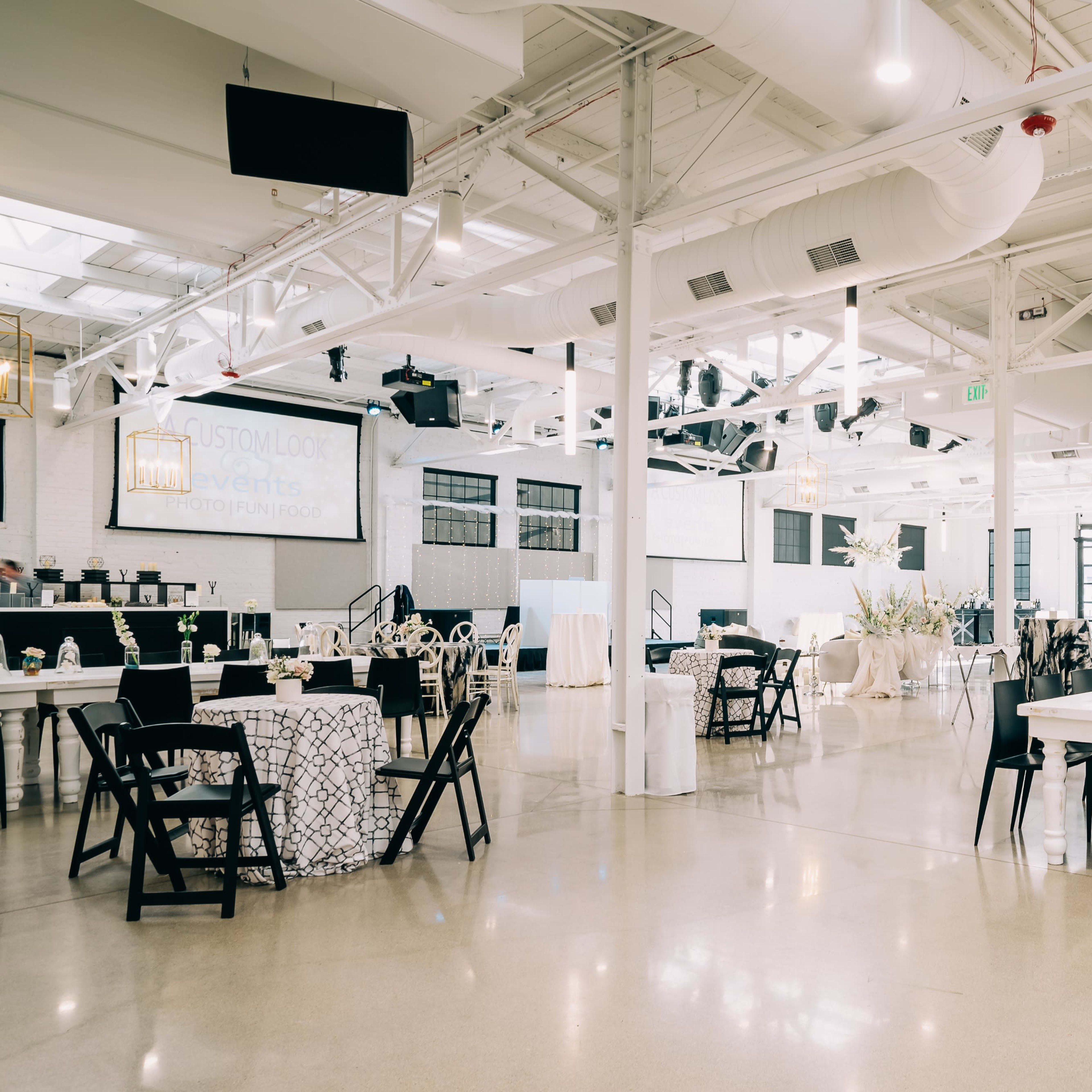 A spacious event venue features long dining tables, black folding chairs, and elegant decor, with large windows and decorative lighting.