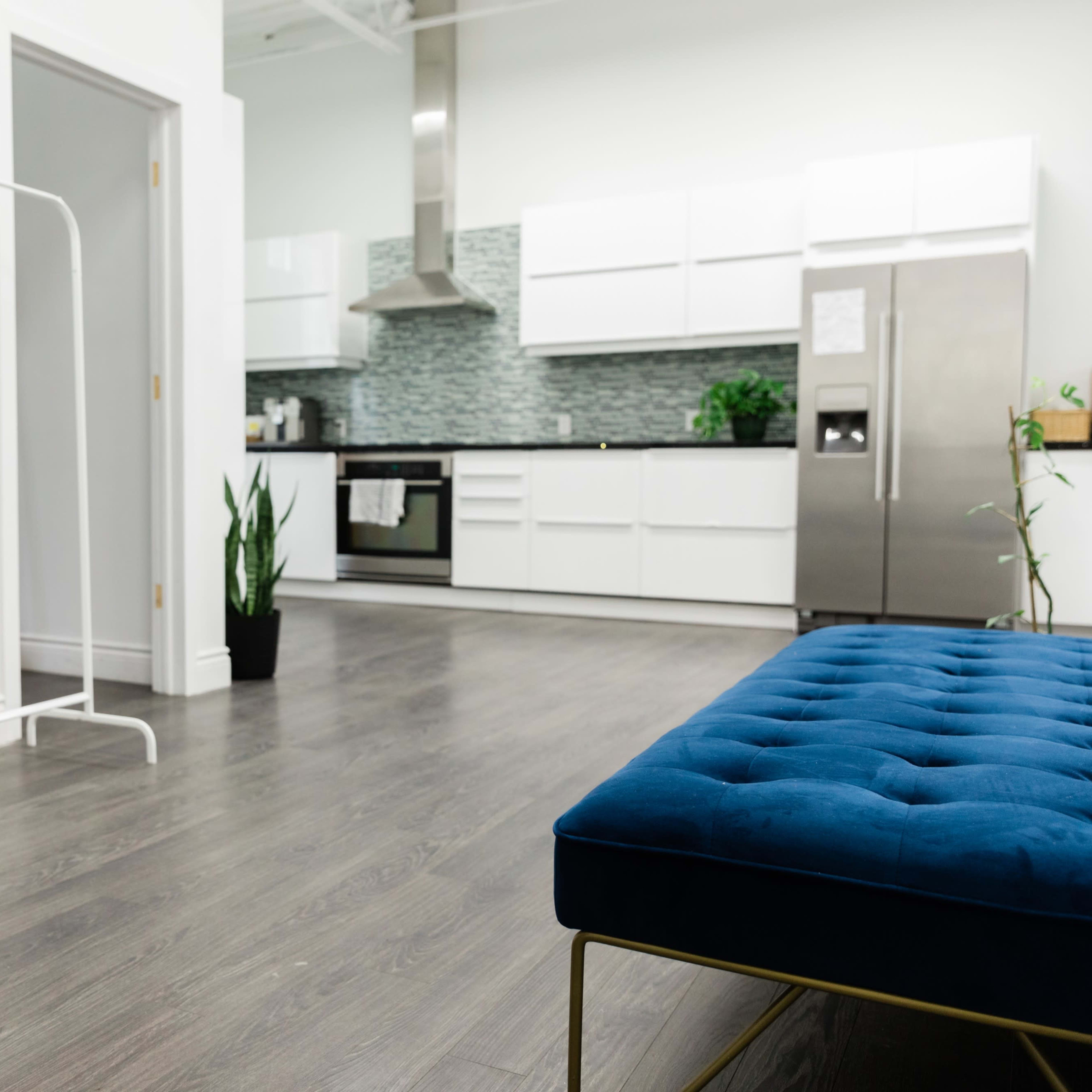 A modern kitchen and living area feature a plush blue bench, a small table with an orange chair, and a clothing rack with plants in the background.