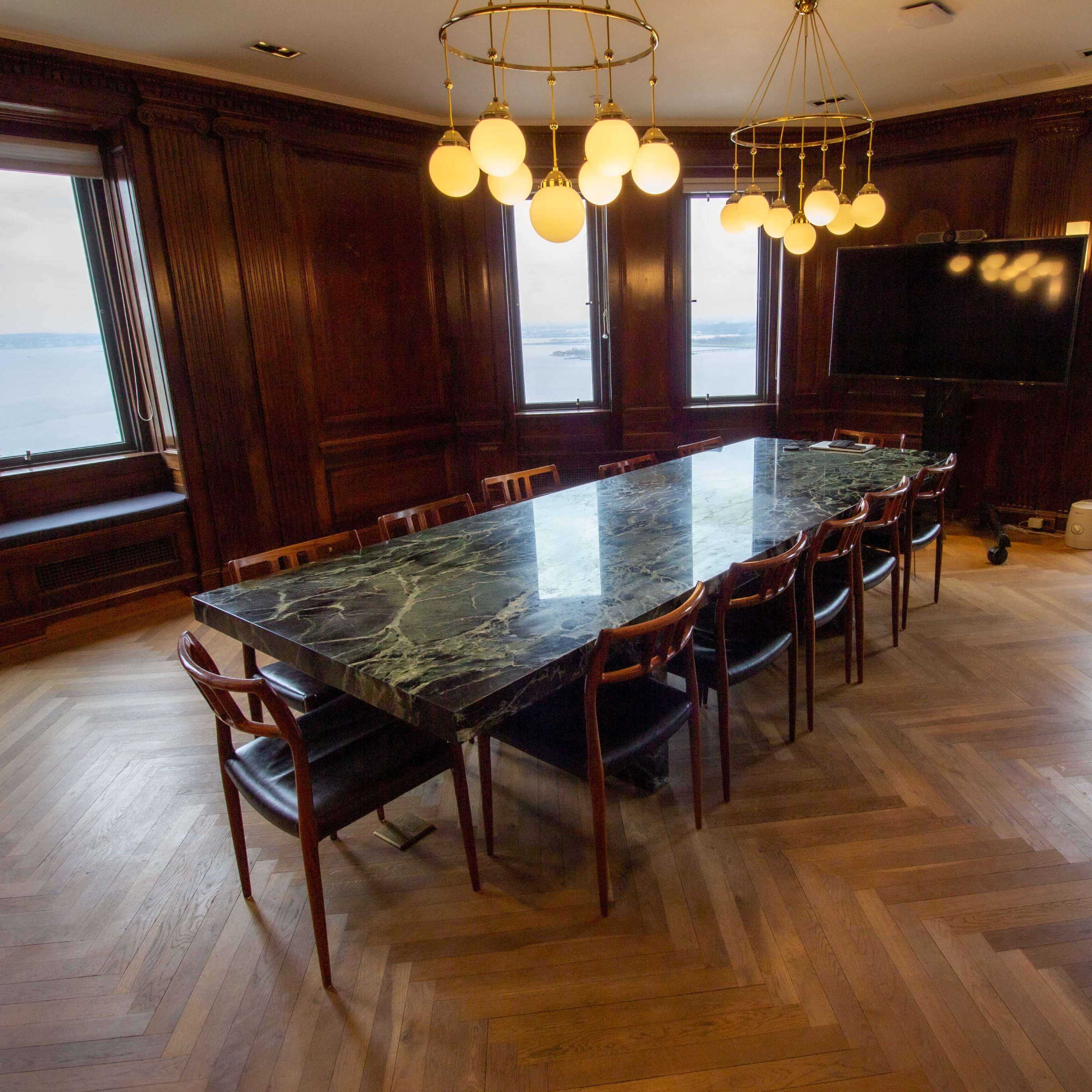 A long rectangular table with a marble top is surrounded by wooden chairs in a conference room featuring large windows with a view of the water.