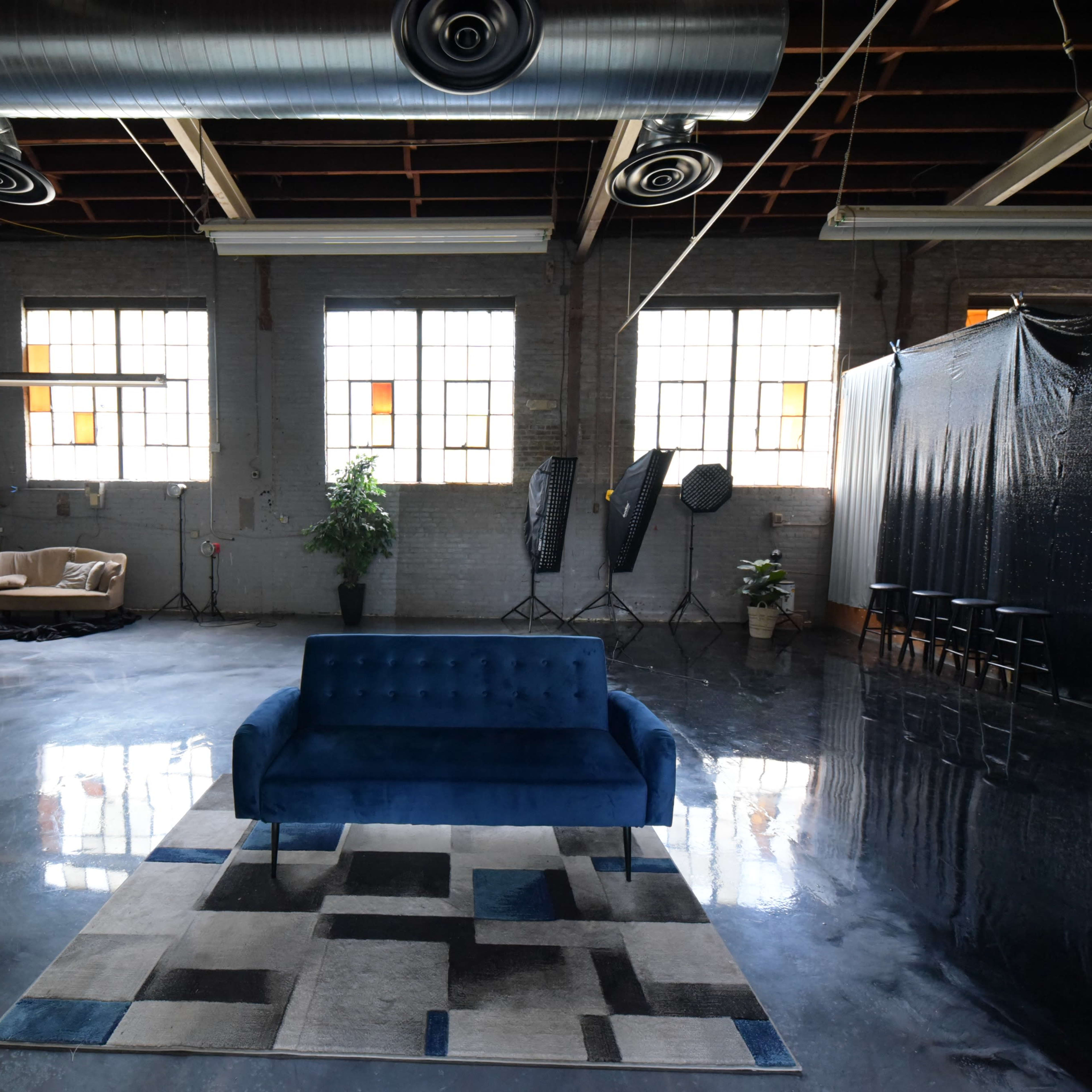 A spacious industrial room with polished concrete floors, large windows, a blue sofa on a patterned rug, and a backdrop set up against one wall.