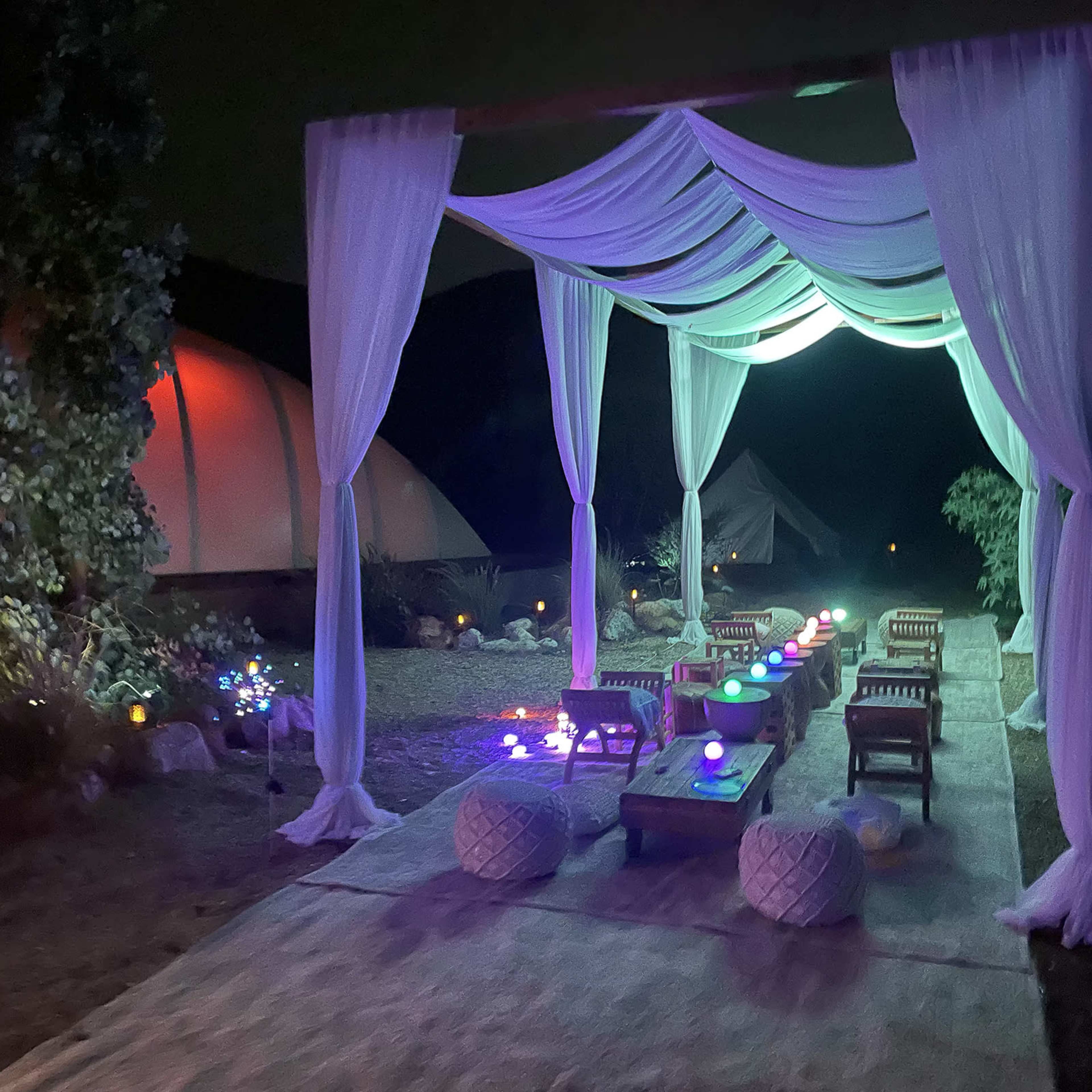A decorated outdoor space features seating under draped fabric illuminated by colored lights at night.