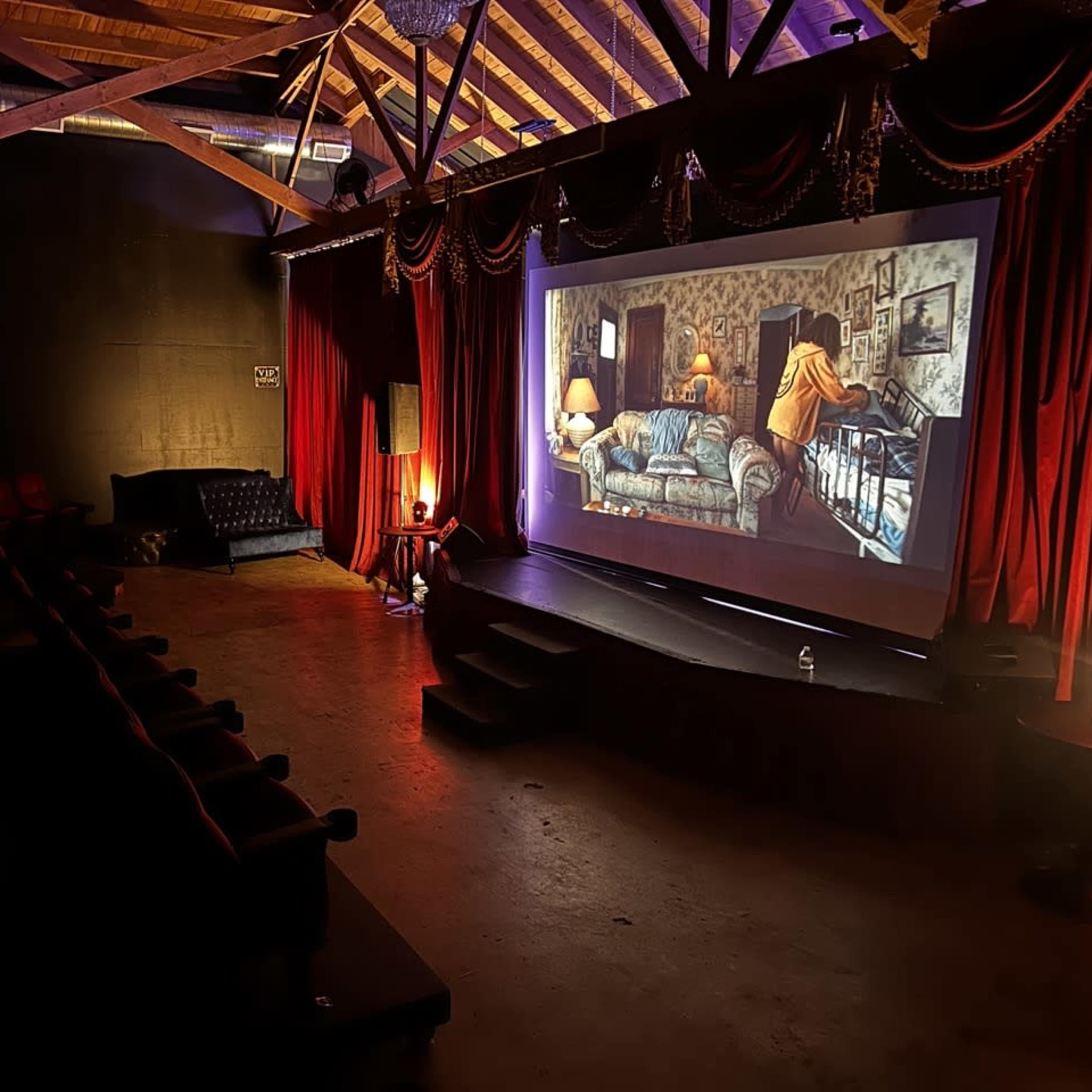 A dimly lit theater contains red velvet curtains and a large screen displaying a scene from a film, with rows of empty seats in front.