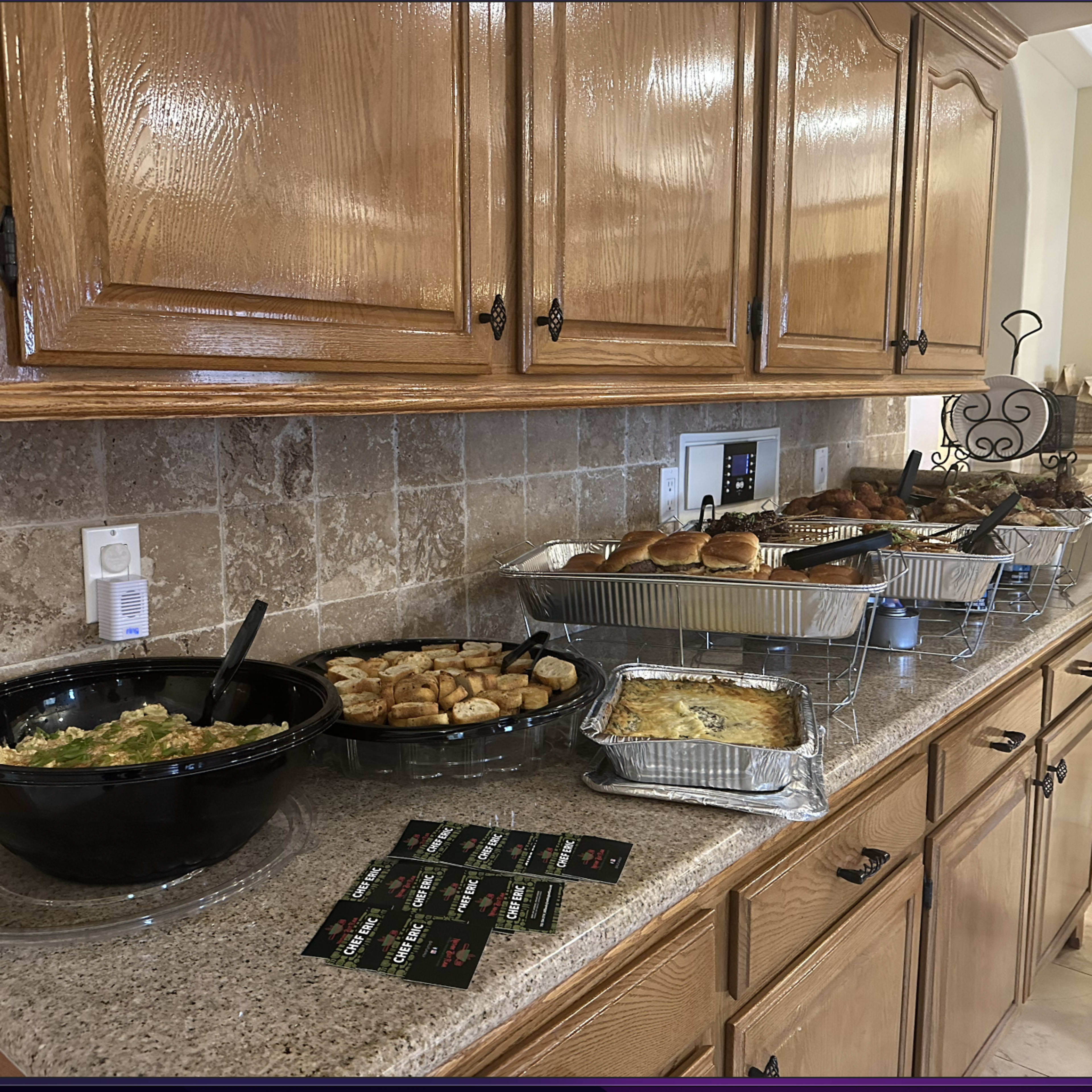 A variety of dishes, including a salad, bread rolls, and a baked casserole, are displayed on a countertop beneath wooden cabinets.