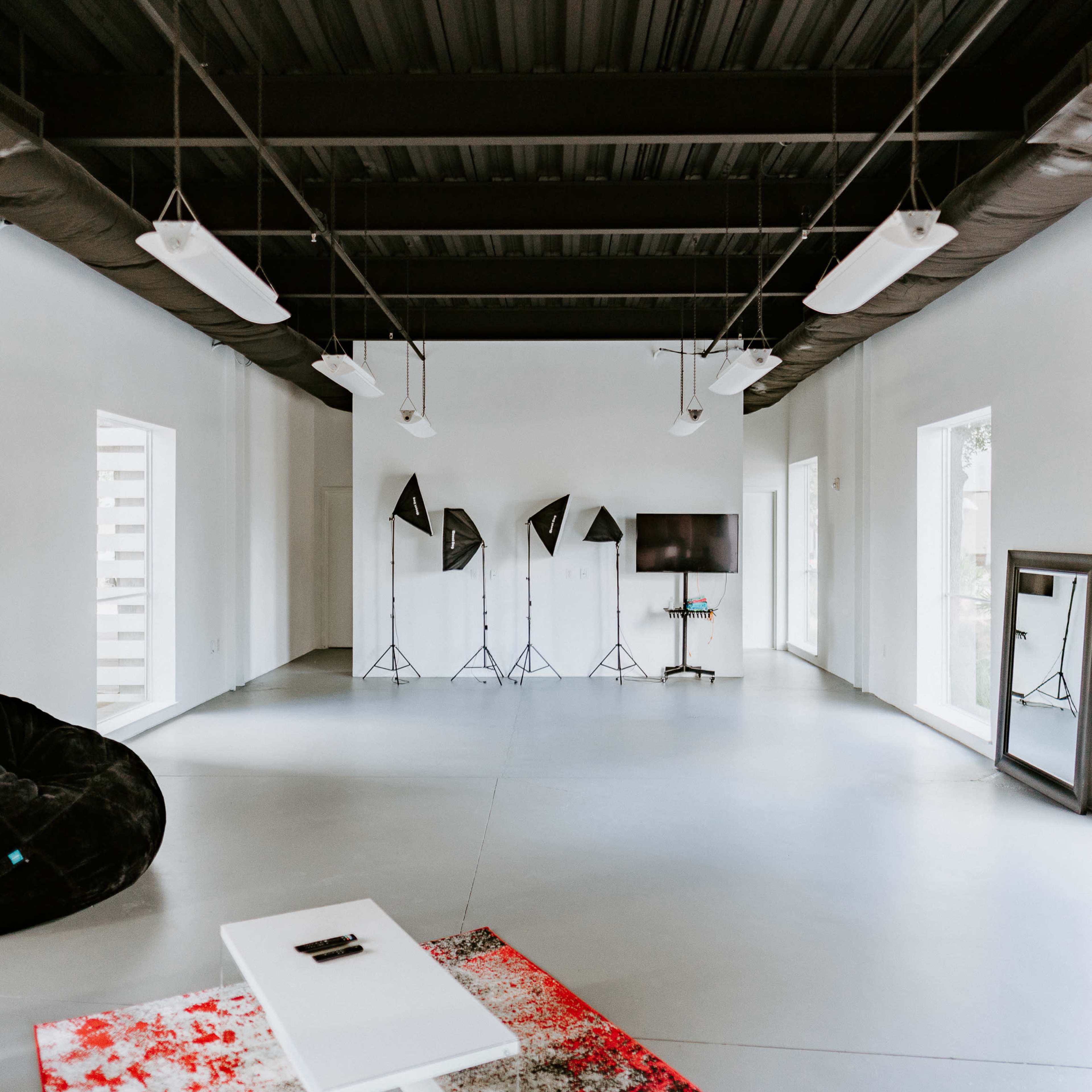 A spacious, minimalist room with gray floors, white walls, and a large red and white rug, featuring several photography studio lights and a screen in the background.