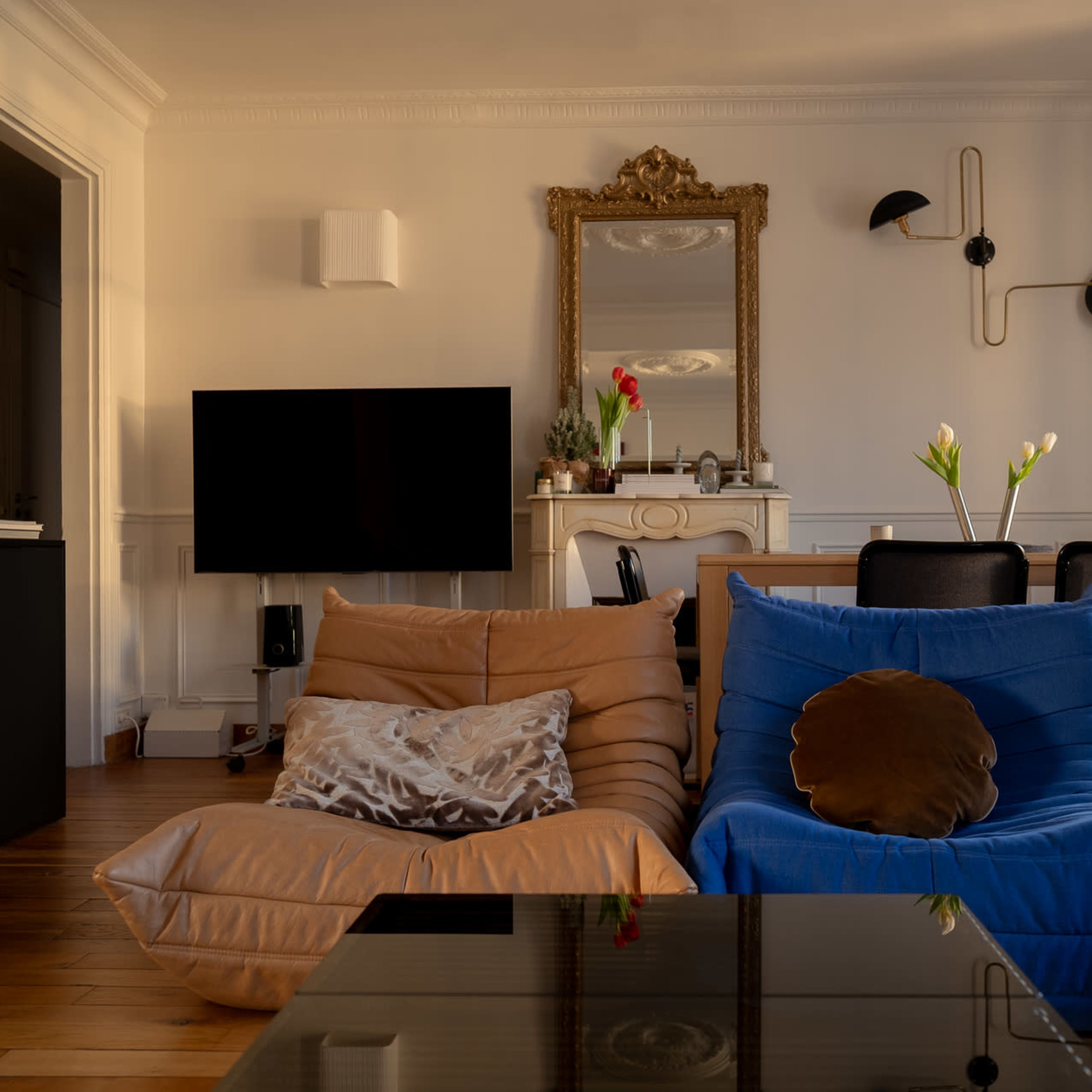 The scene features a living room with a large mirror above a decorative fireplace, two colorful bean bag chairs, a television, and a dining area with a table and chairs.