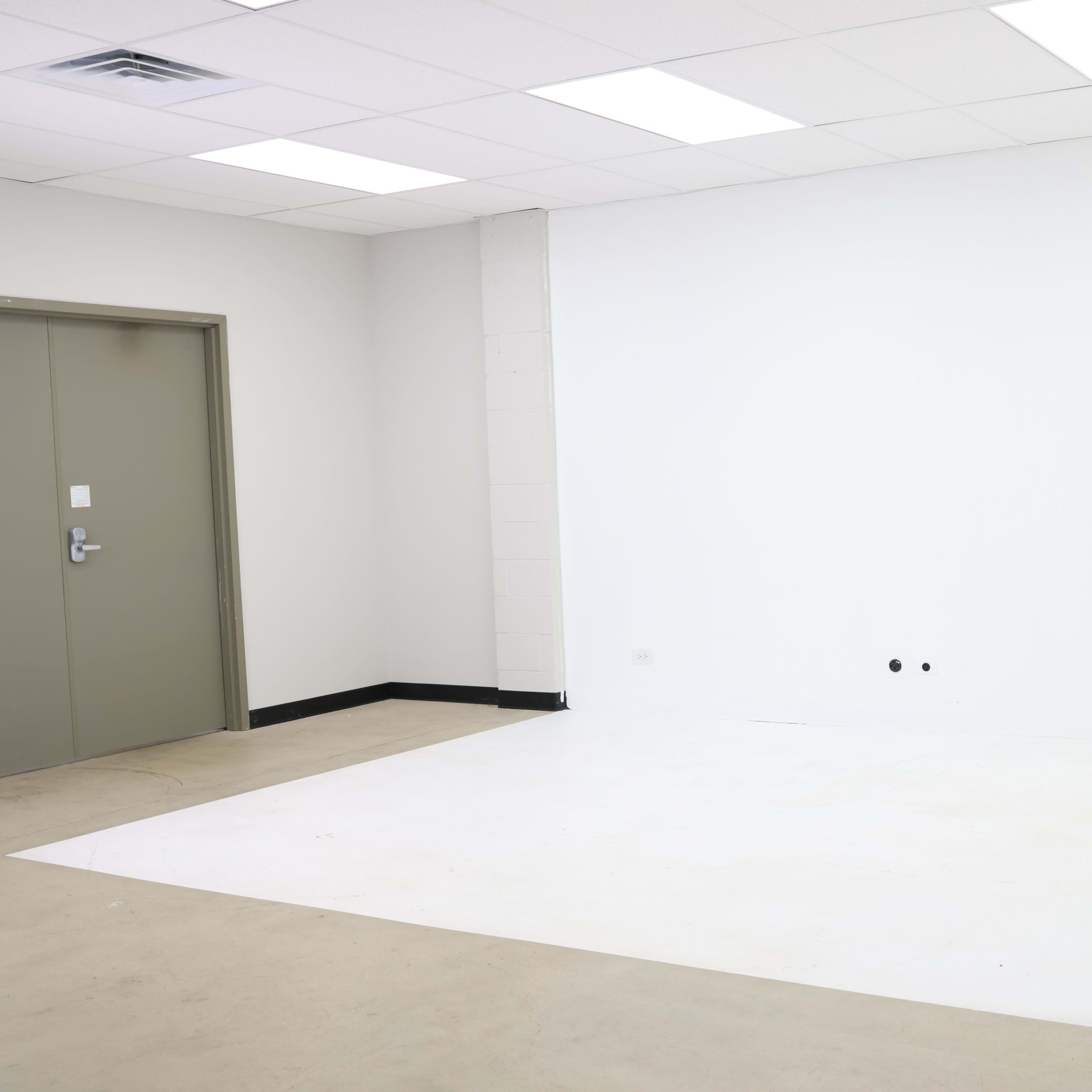 A spacious white room with a light colored floor, a closed door on the left, and an empty wall on the right.