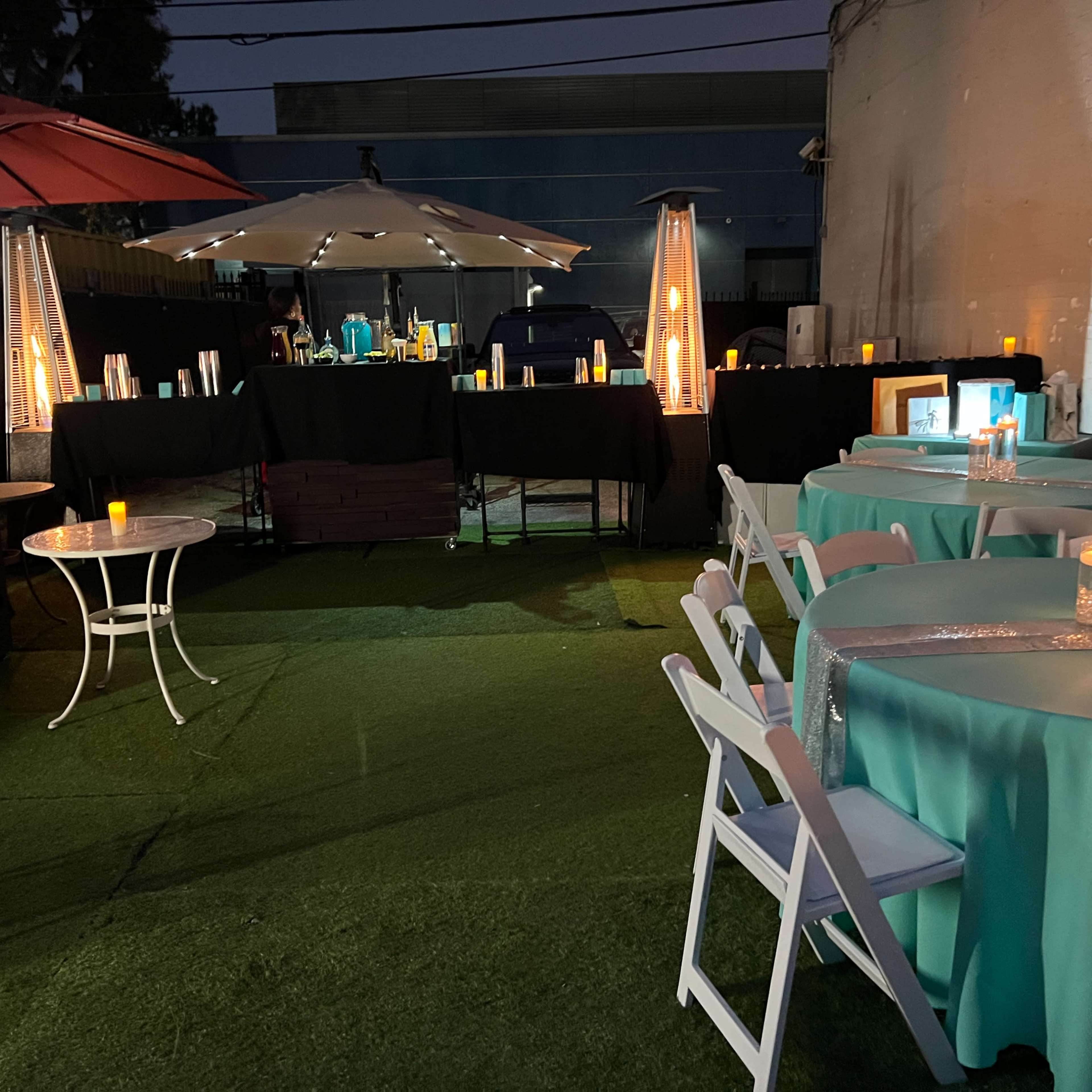An outdoor event area featuring tables with turquoise tablecloths, illuminated candles, and umbrellas, set against a backdrop of a building at dusk.