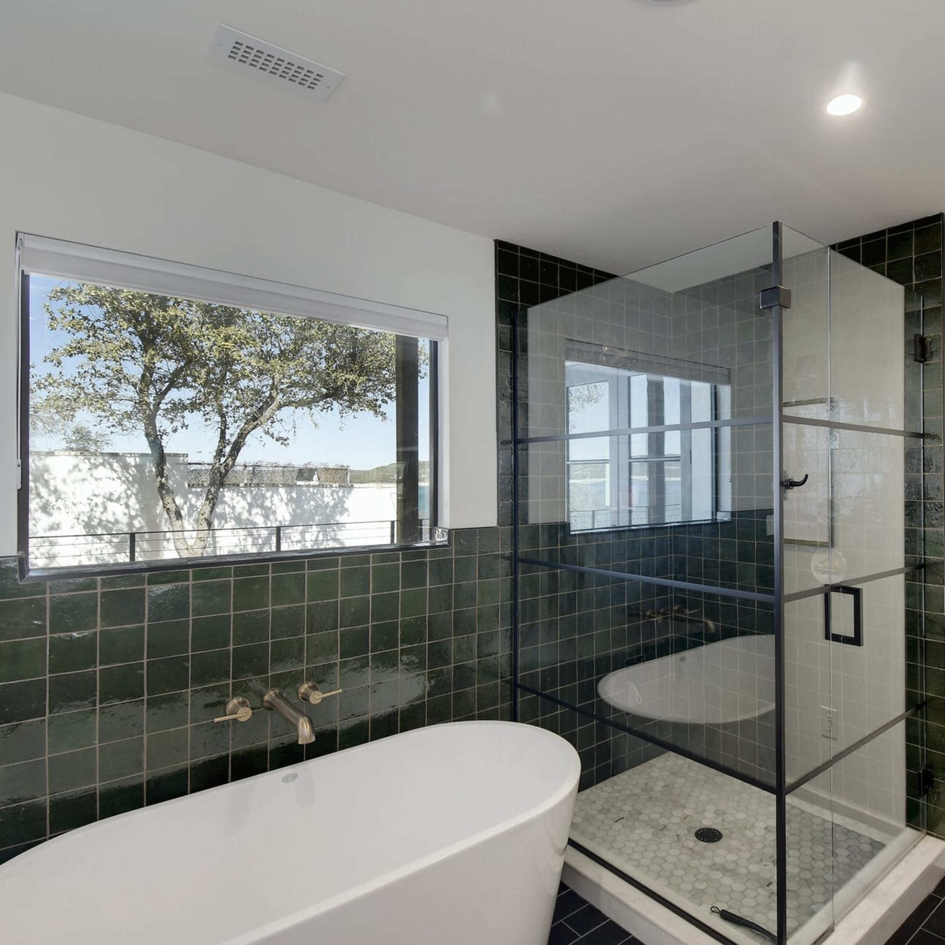 A modern bathroom featuring a glass shower enclosure, a freestanding bathtub, and green tiled walls with a window allowing natural light.