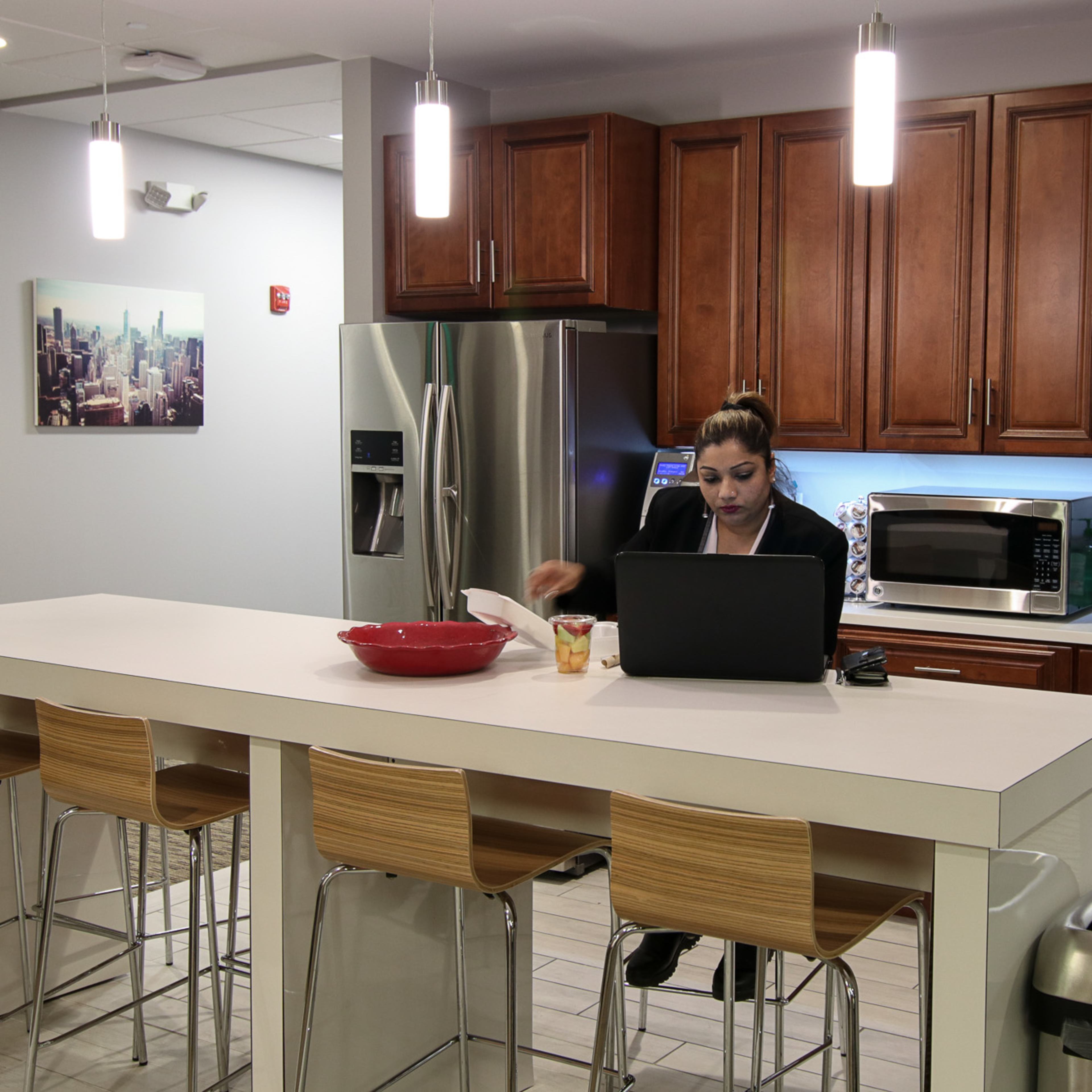 A woman works on a laptop at a kitchen island in a modern kitchen with wooden cabinets and stainless steel appliances.