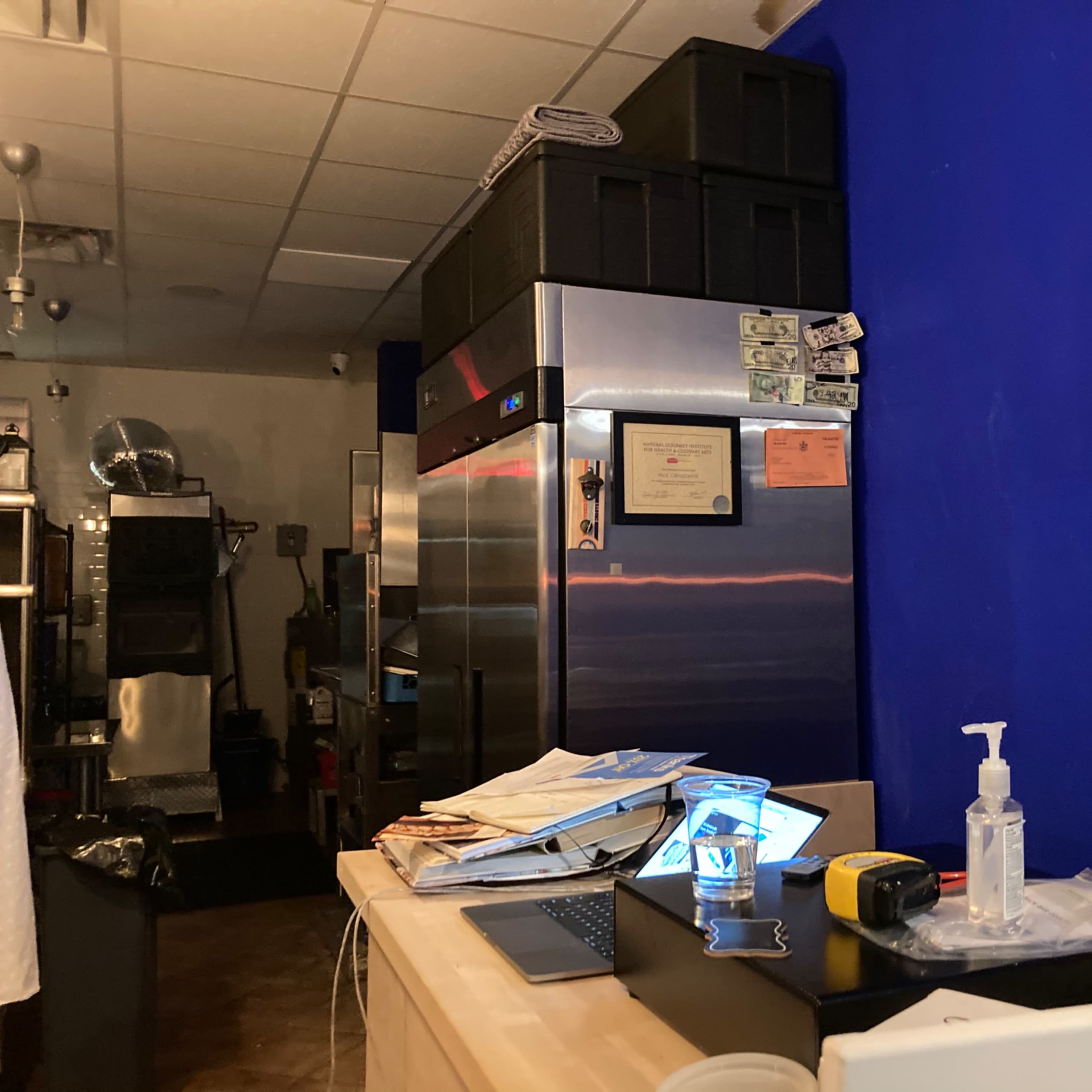 A kitchen counter cluttered with papers, a laptop, and a sanitizer bottle, with a stainless steel refrigerator in the background against a blue wall.