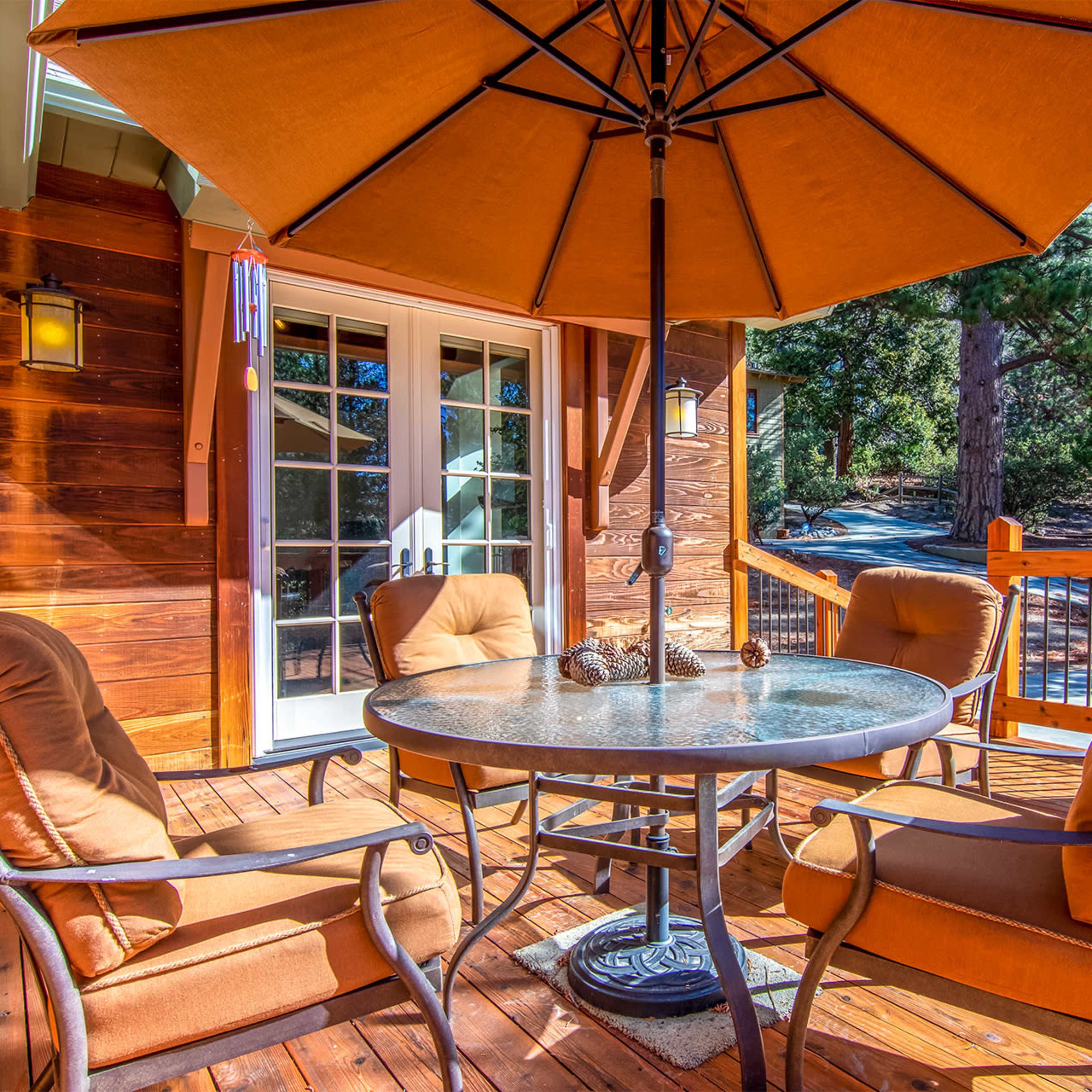 A patio area features a round table with four chairs and a large umbrella, surrounded by wooden decking and forested trees in the background.