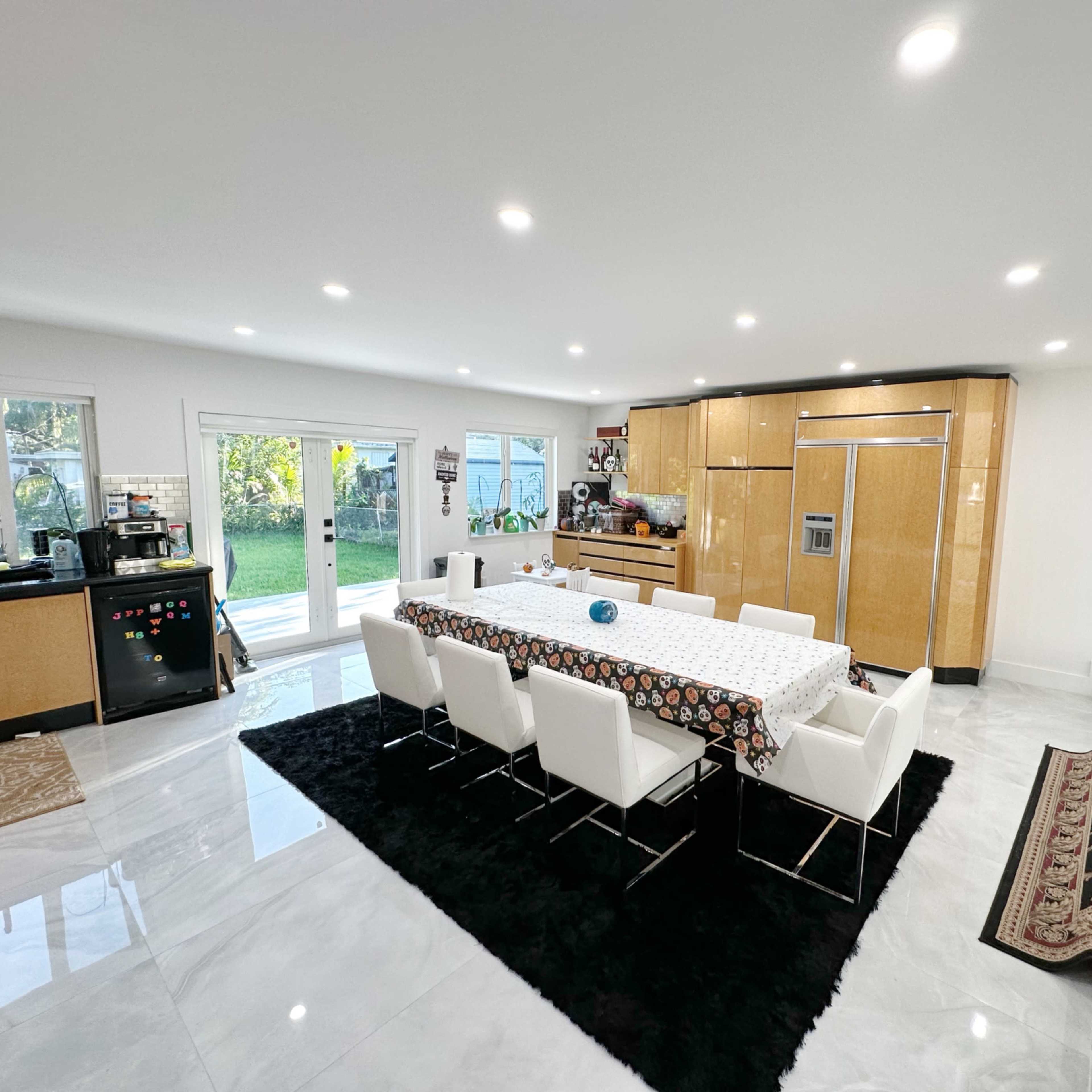 A modern kitchen and dining area features a large table with white chairs, sleek cabinetry, and sliding glass doors leading to a backyard.