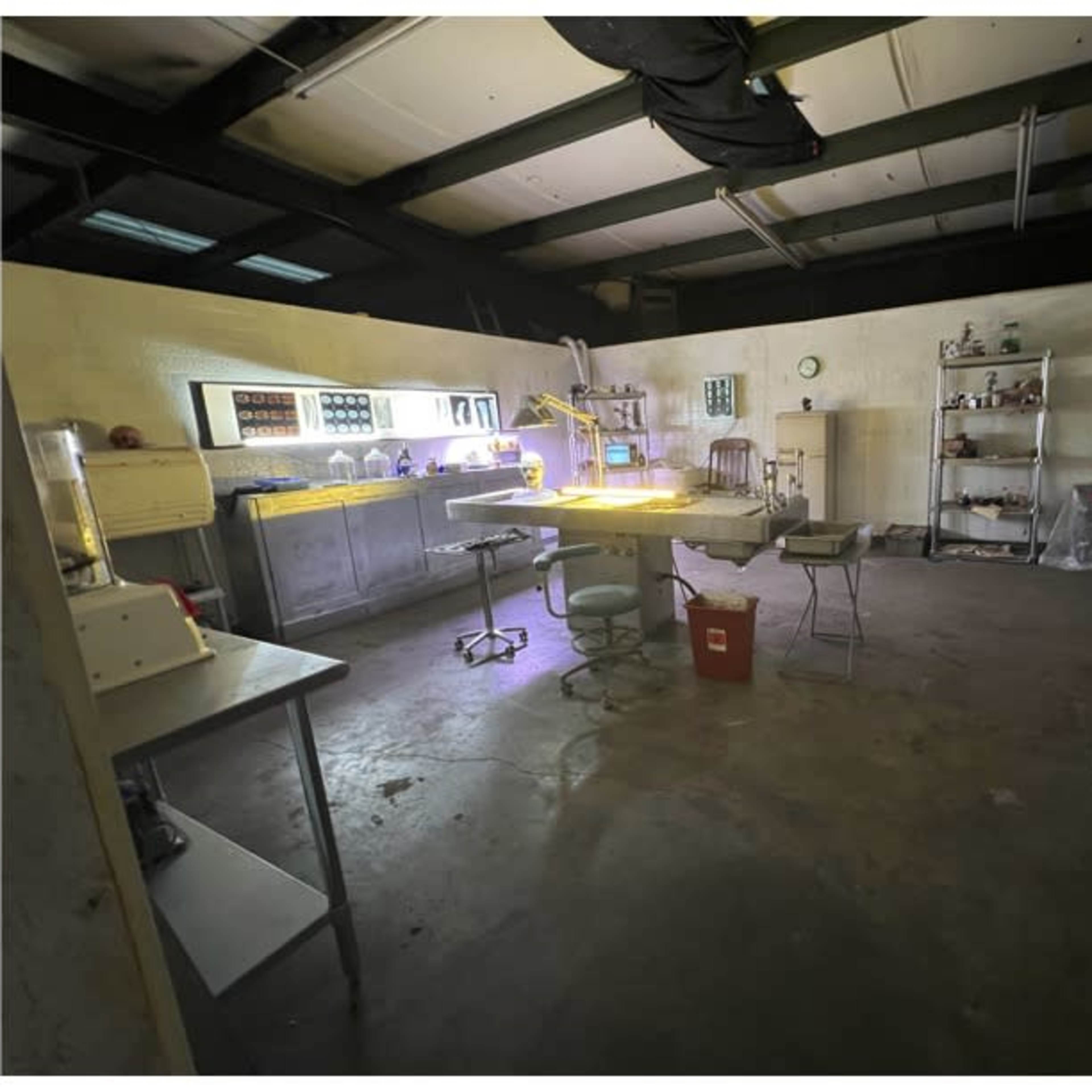 A dimly lit, empty laboratory space featuring a central work table, various instruments, and shelves against the walls.