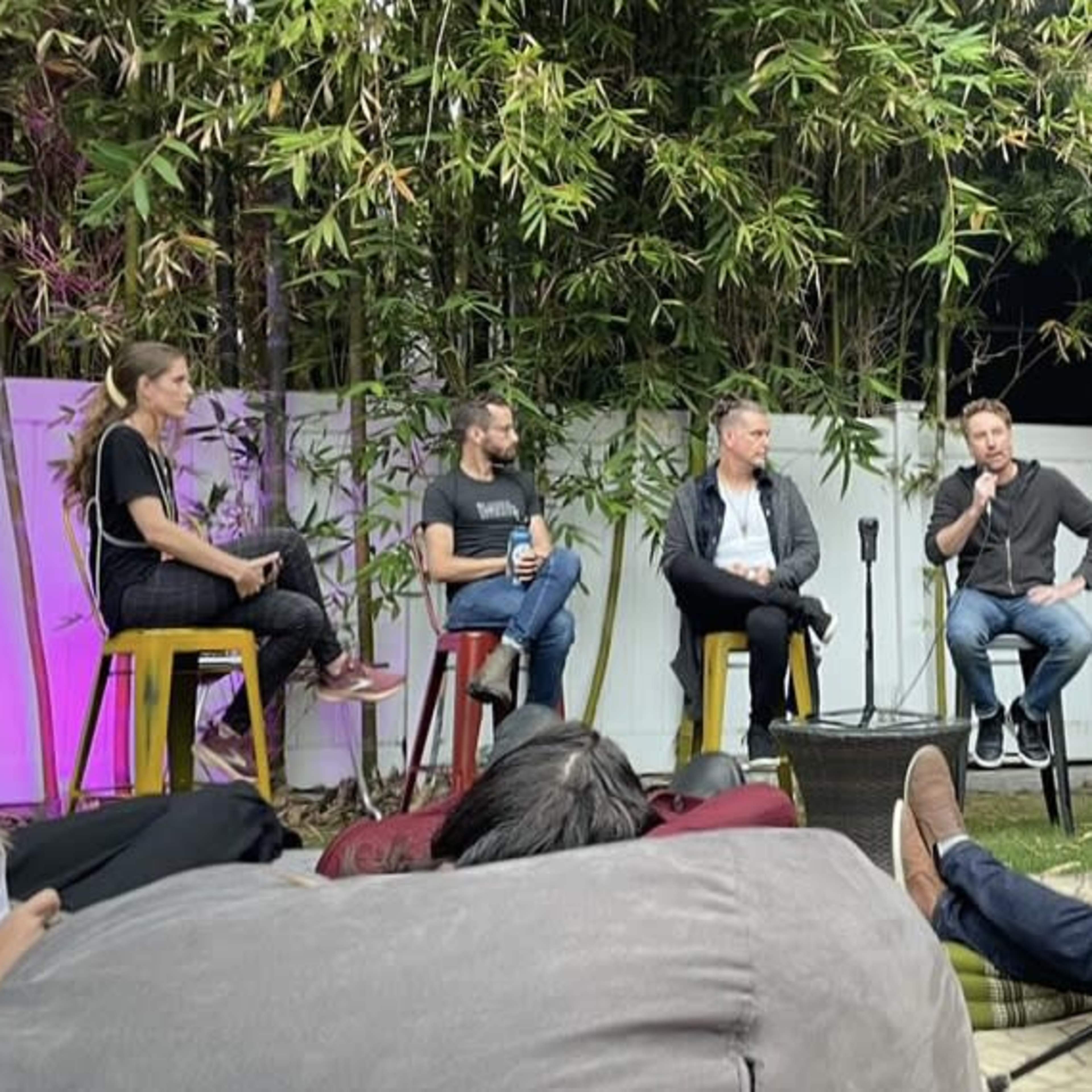 A panel discussion is taking place outdoors with five speakers seated on chairs in front of an audience.