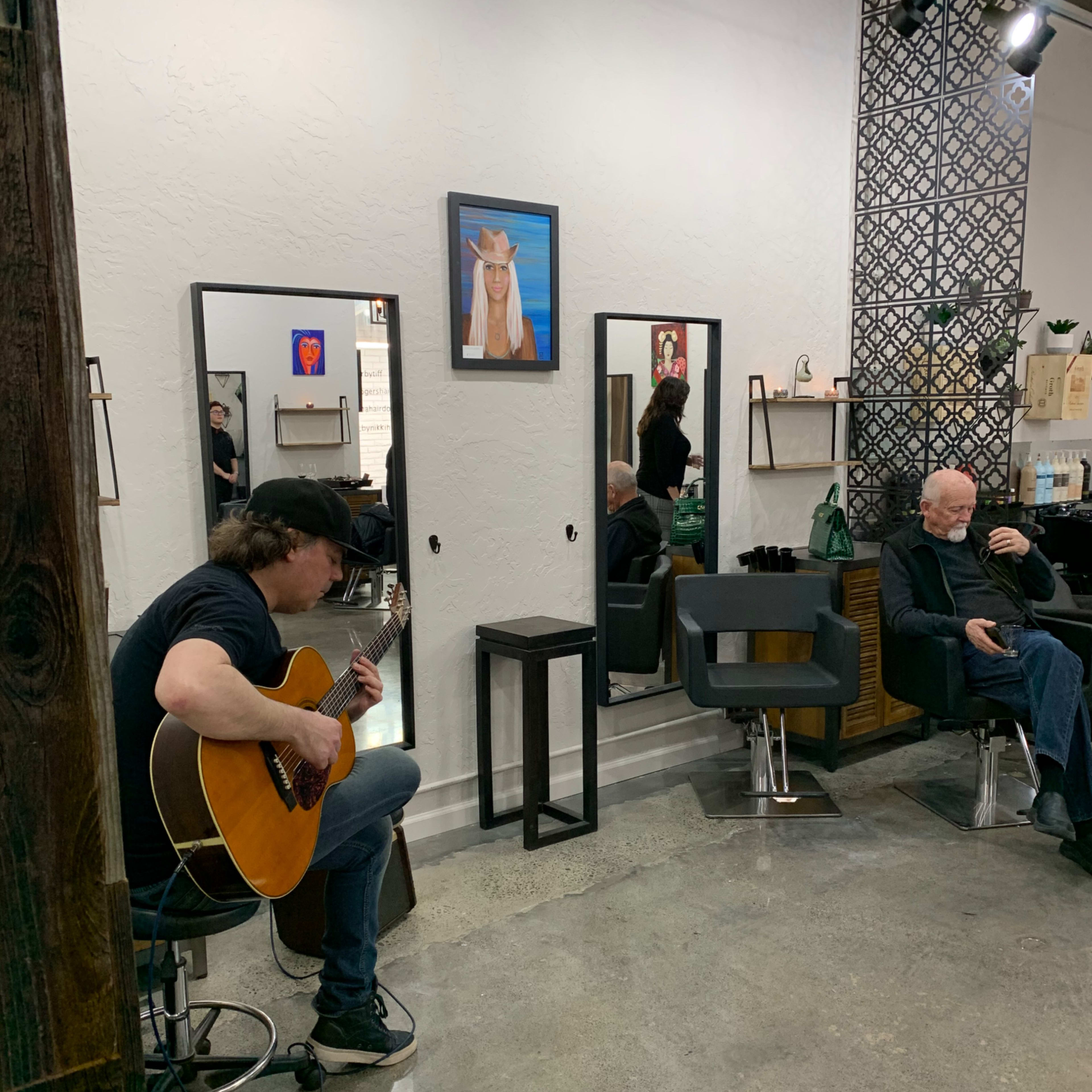 A musician plays guitar in a hair salon while a client sits in a chair getting a haircut.