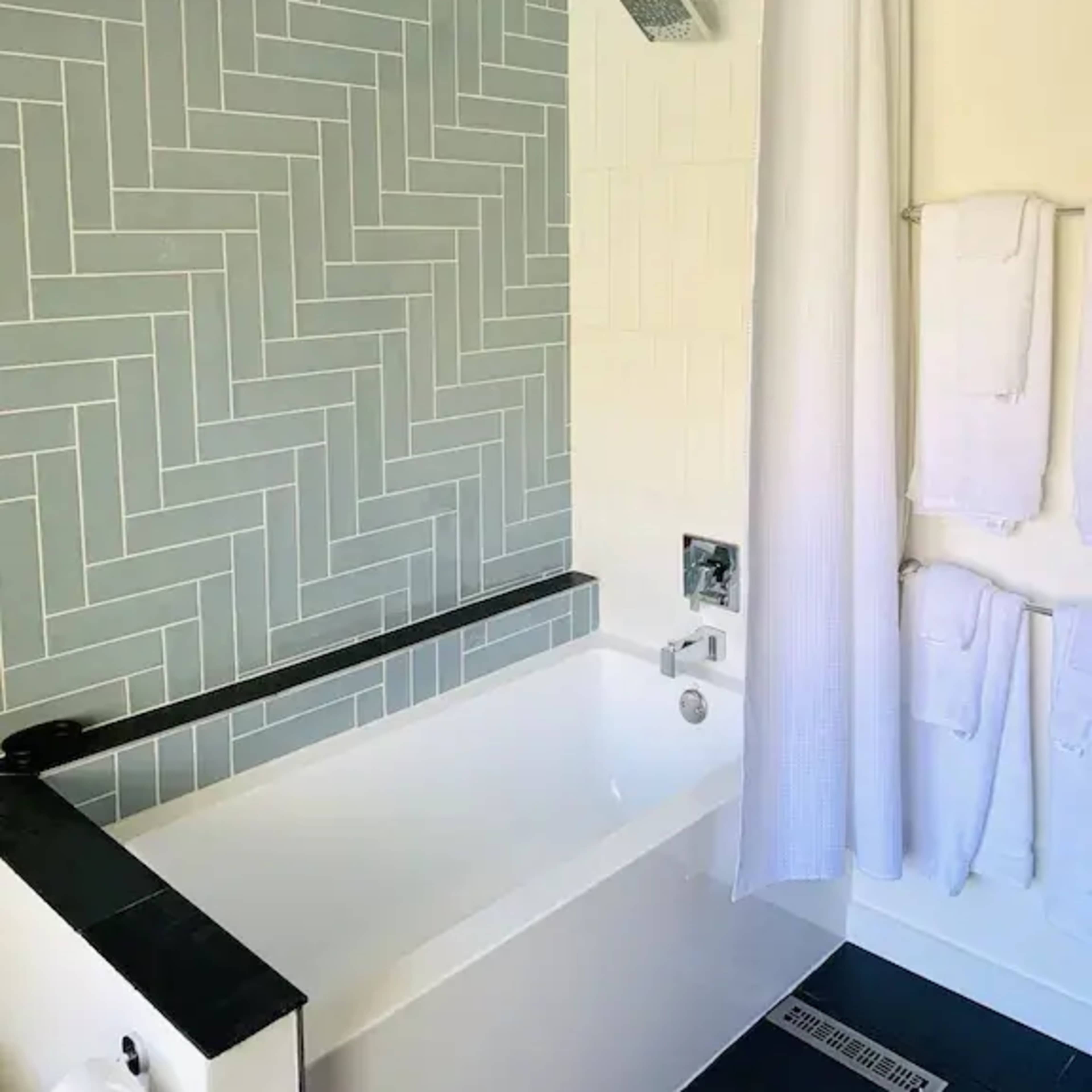 A modern bathroom featuring a bathtub with a herringbone-patterned tile wall, a showerhead, and neatly arranged white towels.